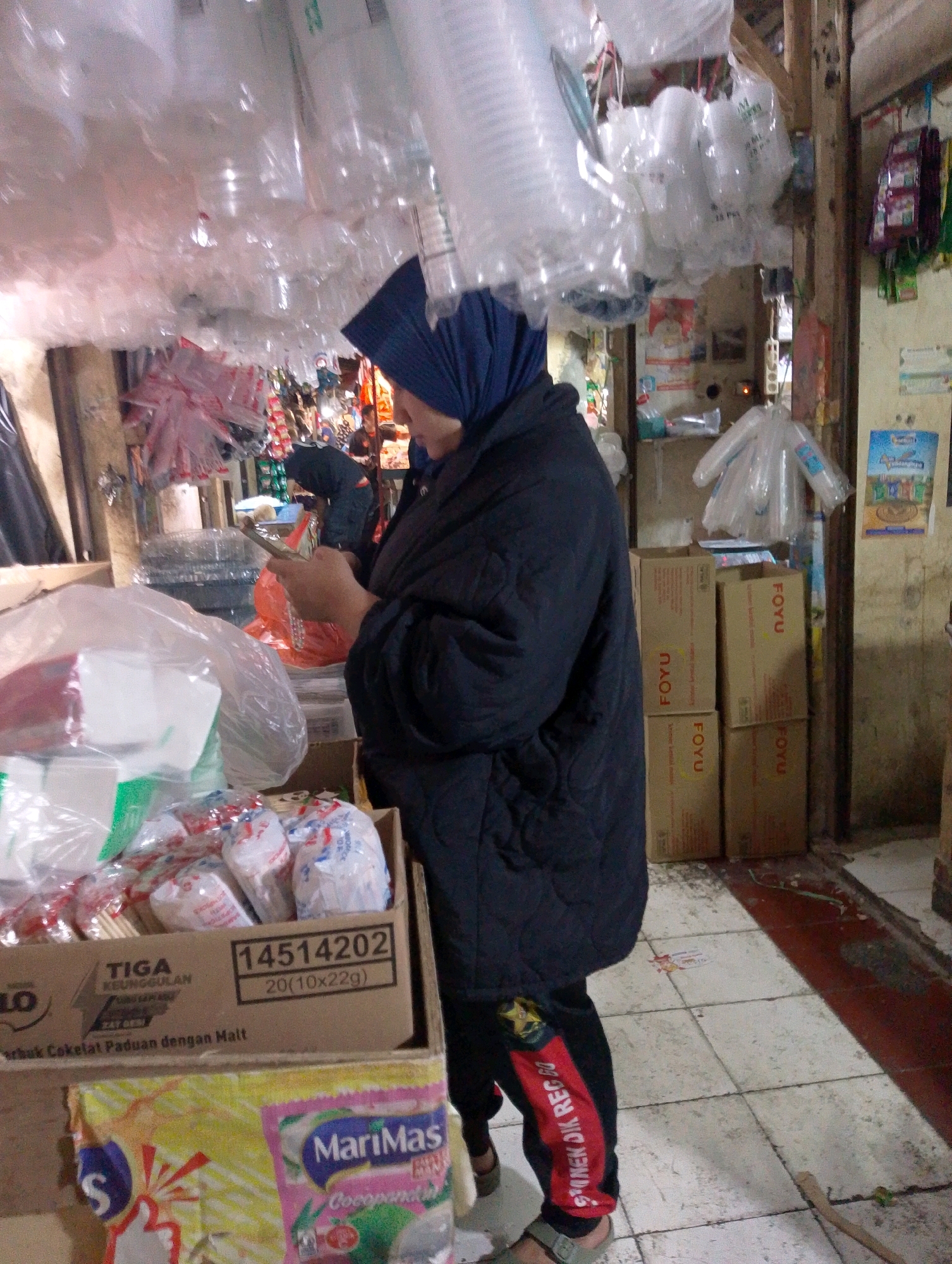 The image depicts a market stall in Lembang, Indonesia, overflowing with various goods. In the foreground, a person wearing a dark blue headscarf and a quilted black jacket is holding a mobile phone and appears to be in conversation or engaged with the device. The stall is laden with plastic cups and containers, some still in their packaging, suspended from the ceiling and stacked on shelves. Boxes of food products are also visible, with labels indicating brands like "TIGA KEUNGGULAN" and "MariMas". The floor is tiled, and a long wooden stick lies on the ground in the lower right corner. The overall impression is of a bustling, somewhat cluttered retail environment typical of a local Indonesian market.