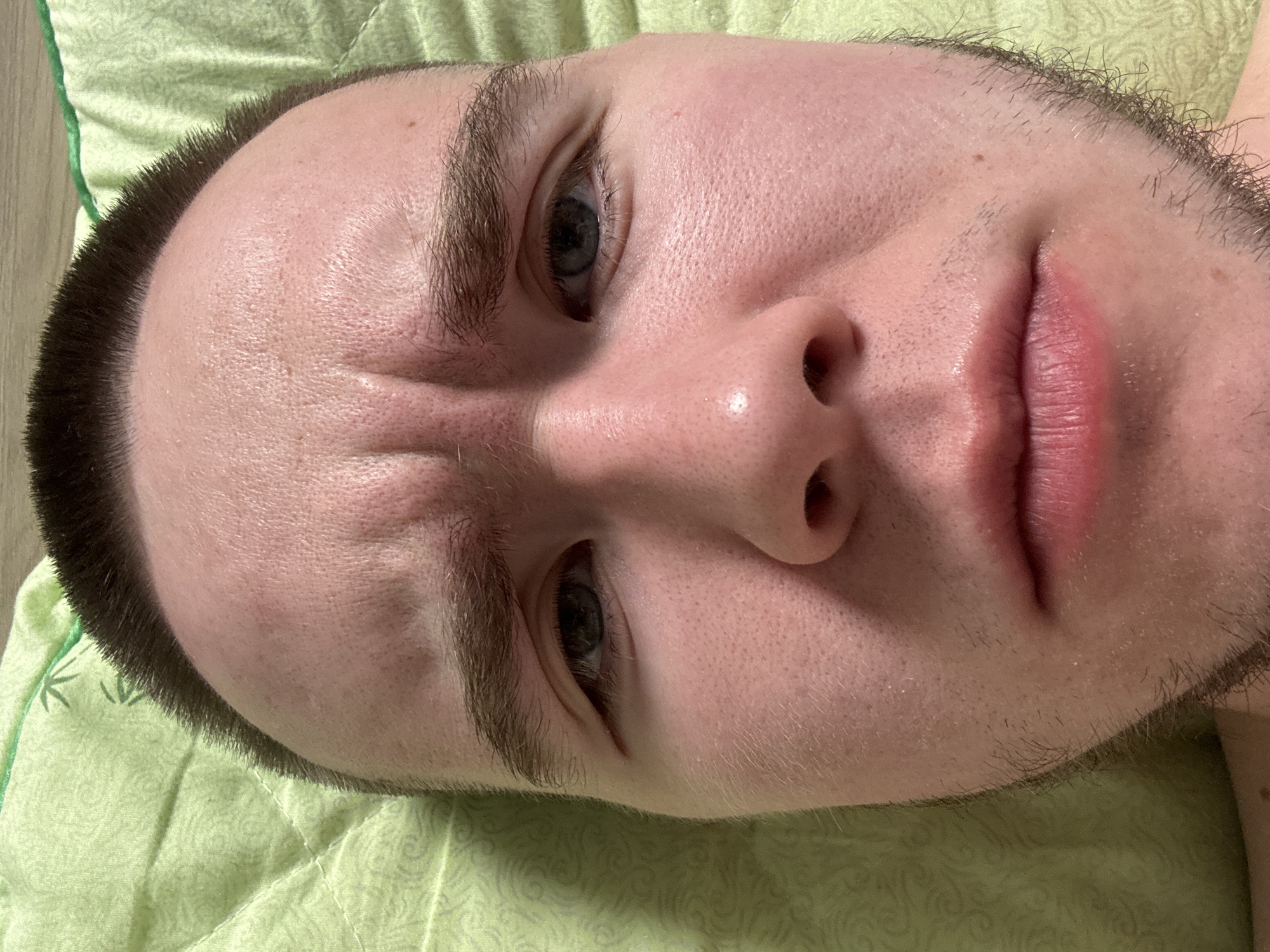 A close-up photograph depicts a person's face, likely male, resting on a light green quilted surface. The subject has fair skin, short dark hair, and light-colored eyes. The facial expression suggests a slight furrowing of the brow. There is no information available to indicate the photograph was taken in Kar'yer, Russia. The only other elements are the quilted fabric visible beneath the face.