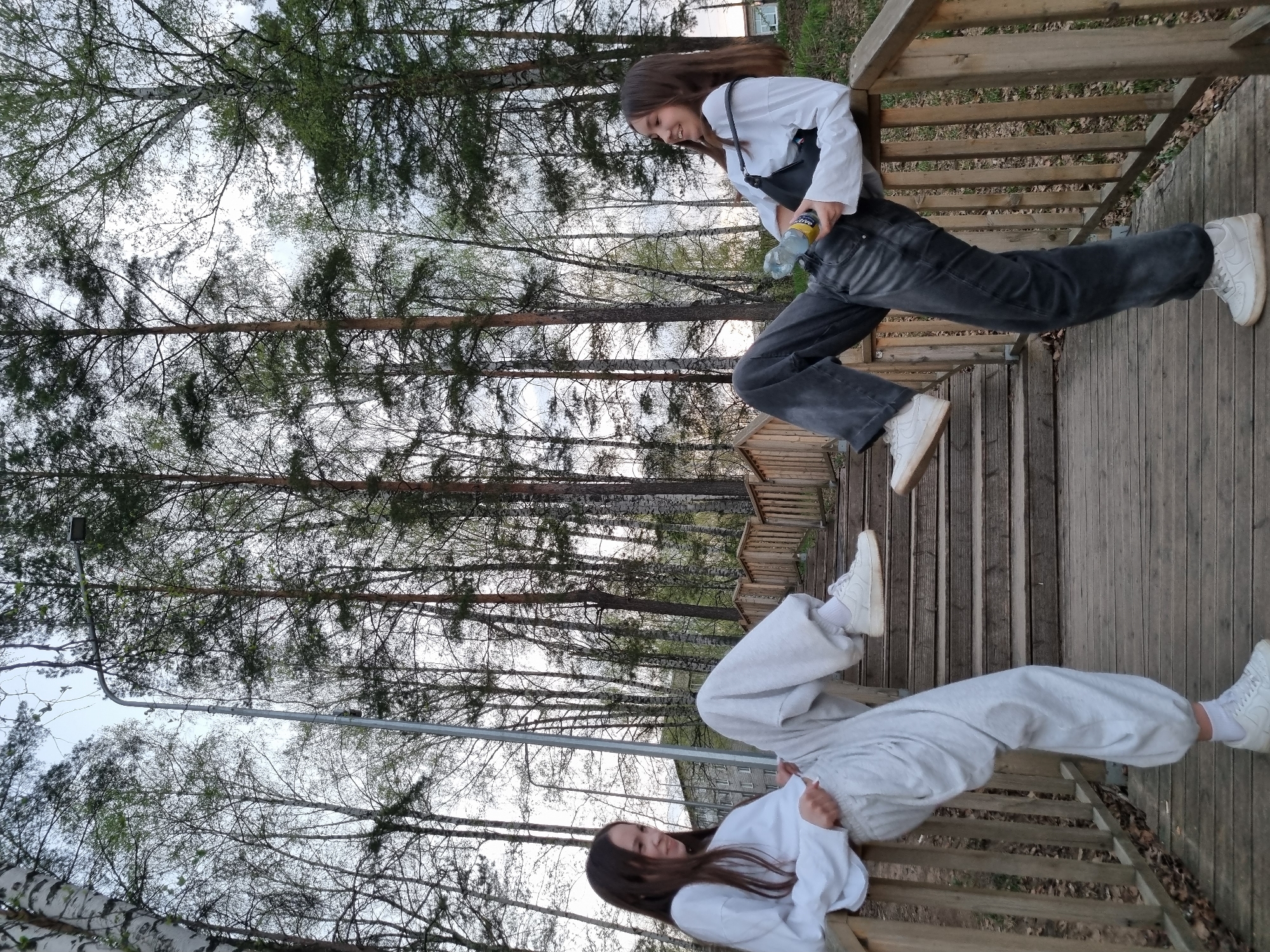 Two young women are positioned on a wooden staircase. One is wearing dark jeans and a white shirt, with a dark cross-body bag, and appears to be holding a bottle. The other is wearing light-colored sweatpants and a matching sweatshirt. The environment includes trees and wooden structures. The location is Chaikovsky, Russia.