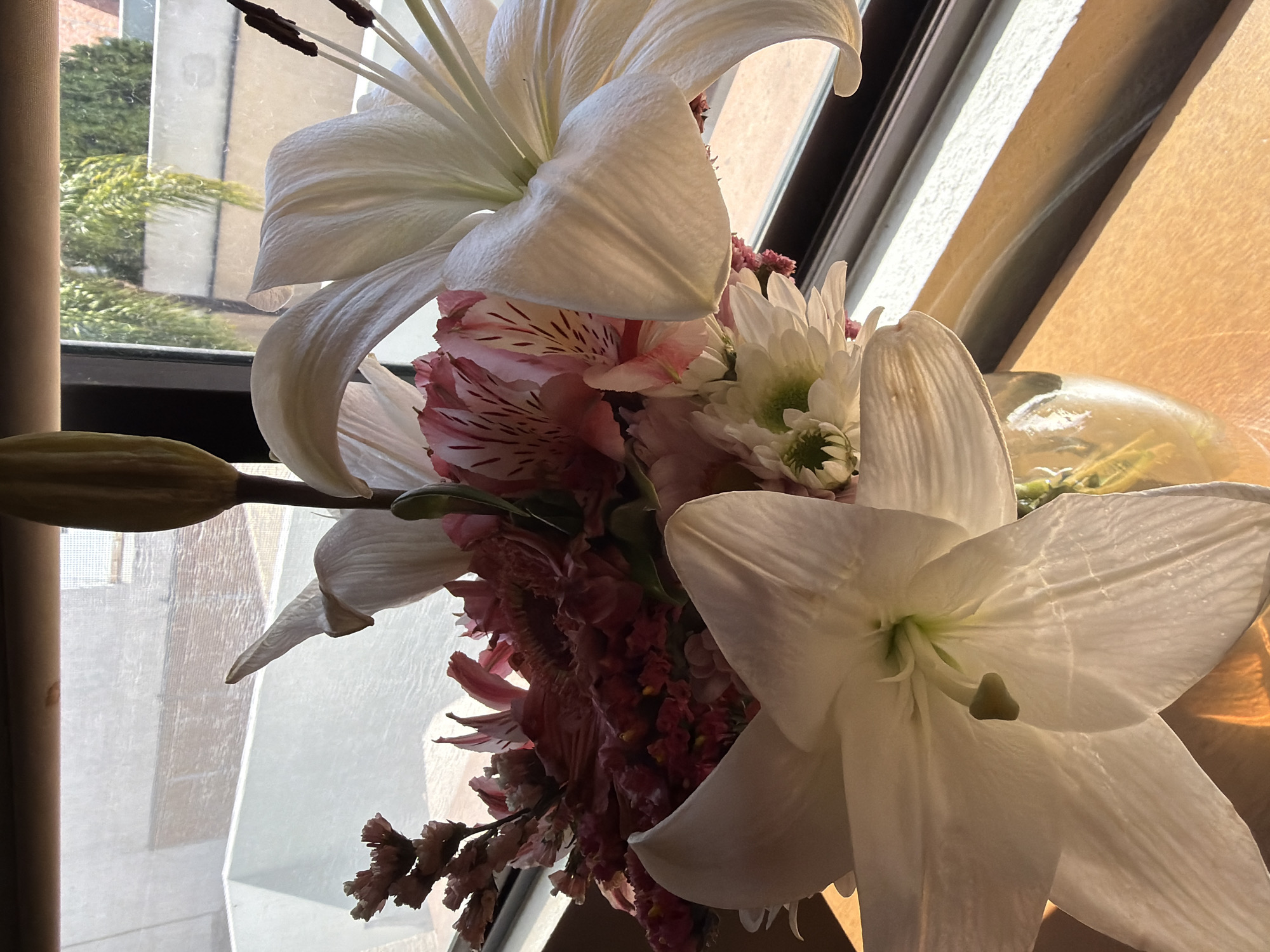 A close-up shot of a bouquet of flowers positioned near a window. The bouquet features white lilies as the dominant floral element, along with pink alstroemeria and white chrysanthemums. One lily is still in bud form. The background shows glimpses of an urban setting through the window, suggesting a cityscape. The location is unknown. The light is bright and possibly natural, casting shadows and highlighting the textures of the flowers.