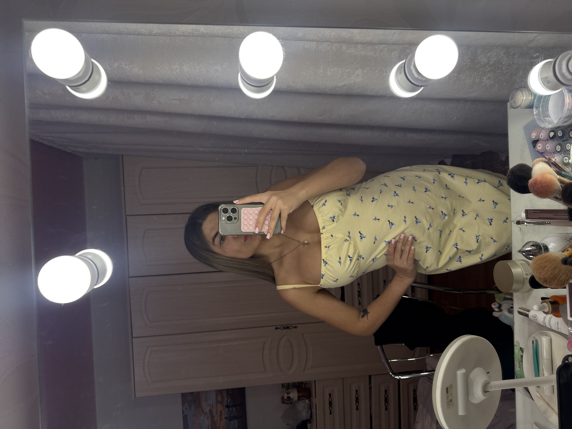 A woman in a yellow floral dress stands in front of a makeup mirror with multiple lights, holding her phone to take a selfie. The mirror is surrounded by makeup and beauty products. The background shows a cabinet with decorative carvings. The location may be Chaikovsky, Russia.