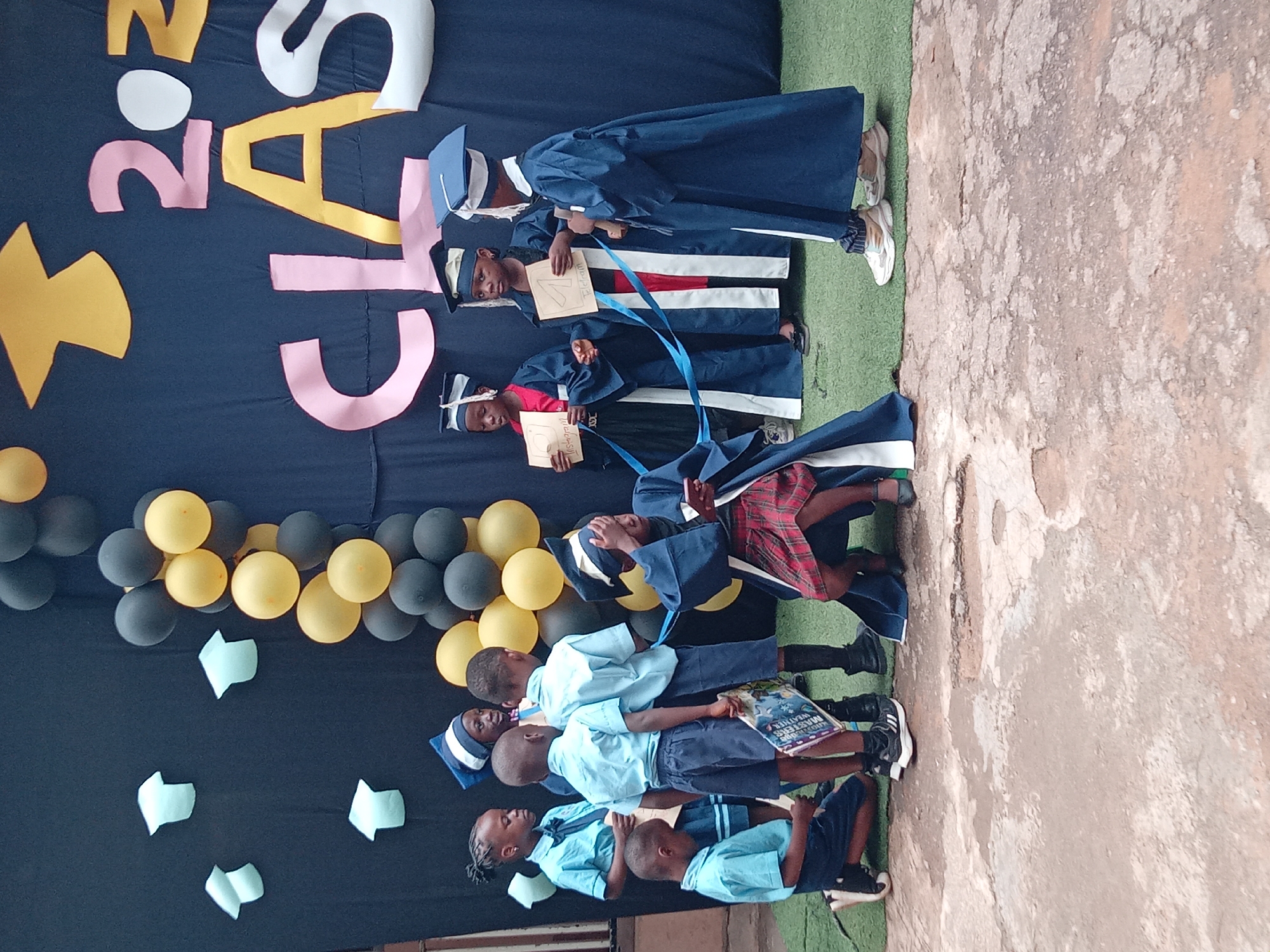 The image depicts a graduation ceremony taking place outdoors, likely in Sabon Tasha, Nigeria. A group of young children are dressed in graduation attire. Some are wearing graduation gowns and caps, while others are in light blue school uniforms. The backdrop features a dark blue cloth decorated with large, colorful cutouts of graduation caps and the numbers "2023" and "CLASS". Balloons in black and yellow are also part of the decoration. Several children are holding up small cards with what appear to be social media platform names written on them, such as "Instagram" and "Telegram." One child in the foreground is kneeling, looking down at a book, while others stand in small groups, some interacting with each other, others looking at the camera or the graduation backdrop. The ground appears to be a mix of rough concrete and green artificial turf.