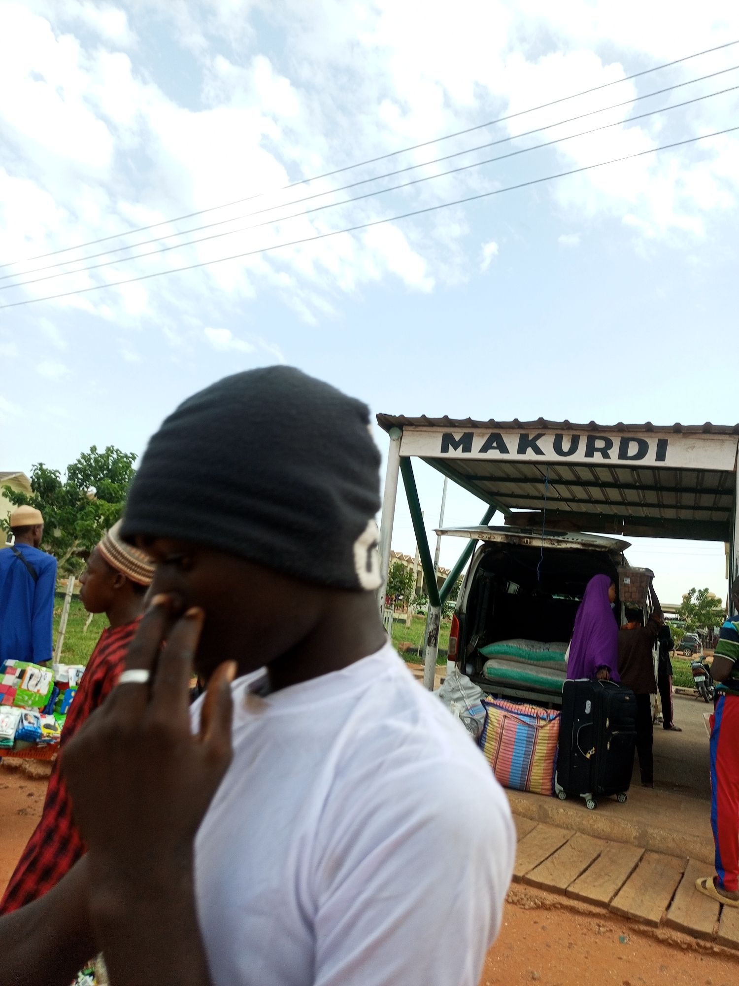 The image depicts an outdoor scene under a partly cloudy sky. A sign above a covered area reads "MAKURDI". In the foreground, a person wearing a black beanie and a white t-shirt is partially visible, with their hand near their face. To the left, other individuals are present, including one wearing a blue garment and another in a red and black checkered shirt. A van with its rear doors open is parked under the covered area, with bags loaded inside. Luggage, including a large colorful duffel bag and a black suitcase, sits beside the van. The ground is covered in red dirt and some wooden planks. In the background, trees and buildings are visible, suggesting an urban or semi-urban environment. The presence of the sign "MAKURDI" indicates the location is likely a transportation hub or a place associated with the city of Makurdi, which is located in Benue State, Nigeria. While the prompt mentions Gombe, Nigeria, the signage specifically points to Makurdi.