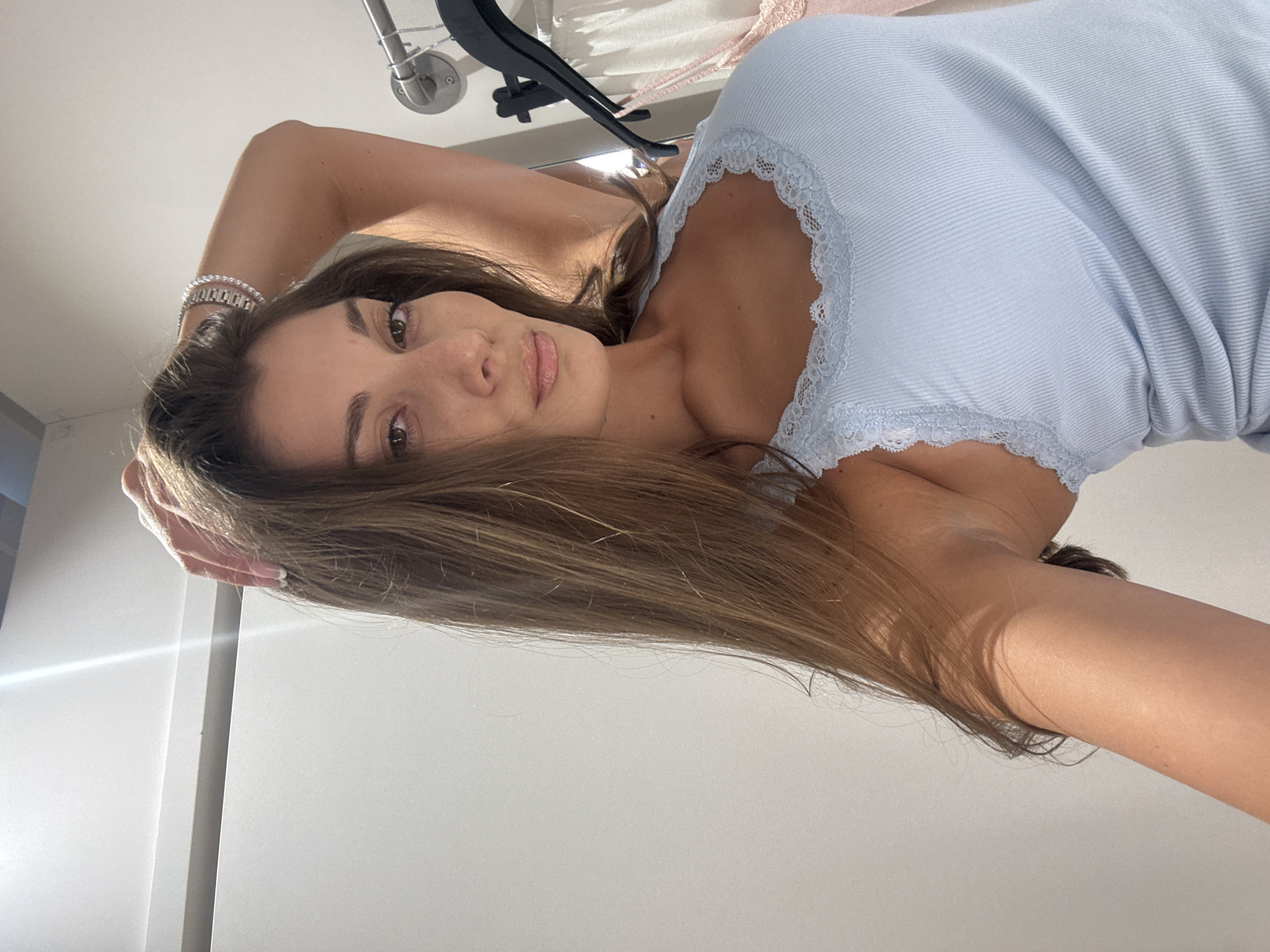 A woman with long brown hair and light skin is lying on a surface, facing the camera. She is wearing a light blue ribbed tank top with a white lace trim. A silver bracelet is visible on her left wrist. A clothing rack with clothes on hangers is in the background. The setting appears to be an indoor space with a white wall. The location is not discernible as Burgas, Bulgaria from the given image.