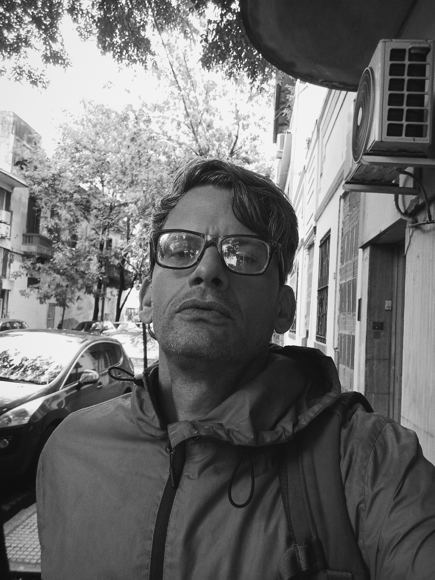 A close-up, black and white photograph depicts a man wearing glasses and a hooded jacket, facing the camera. He is positioned on a city street. Behind him to the left is a parked car and trees line the street. To the right, a building wall features windows and an air conditioning unit. The photo appears to be taken in Buenos Aires, Argentina. The architectural style of the buildings suggests a European influence common in the city.