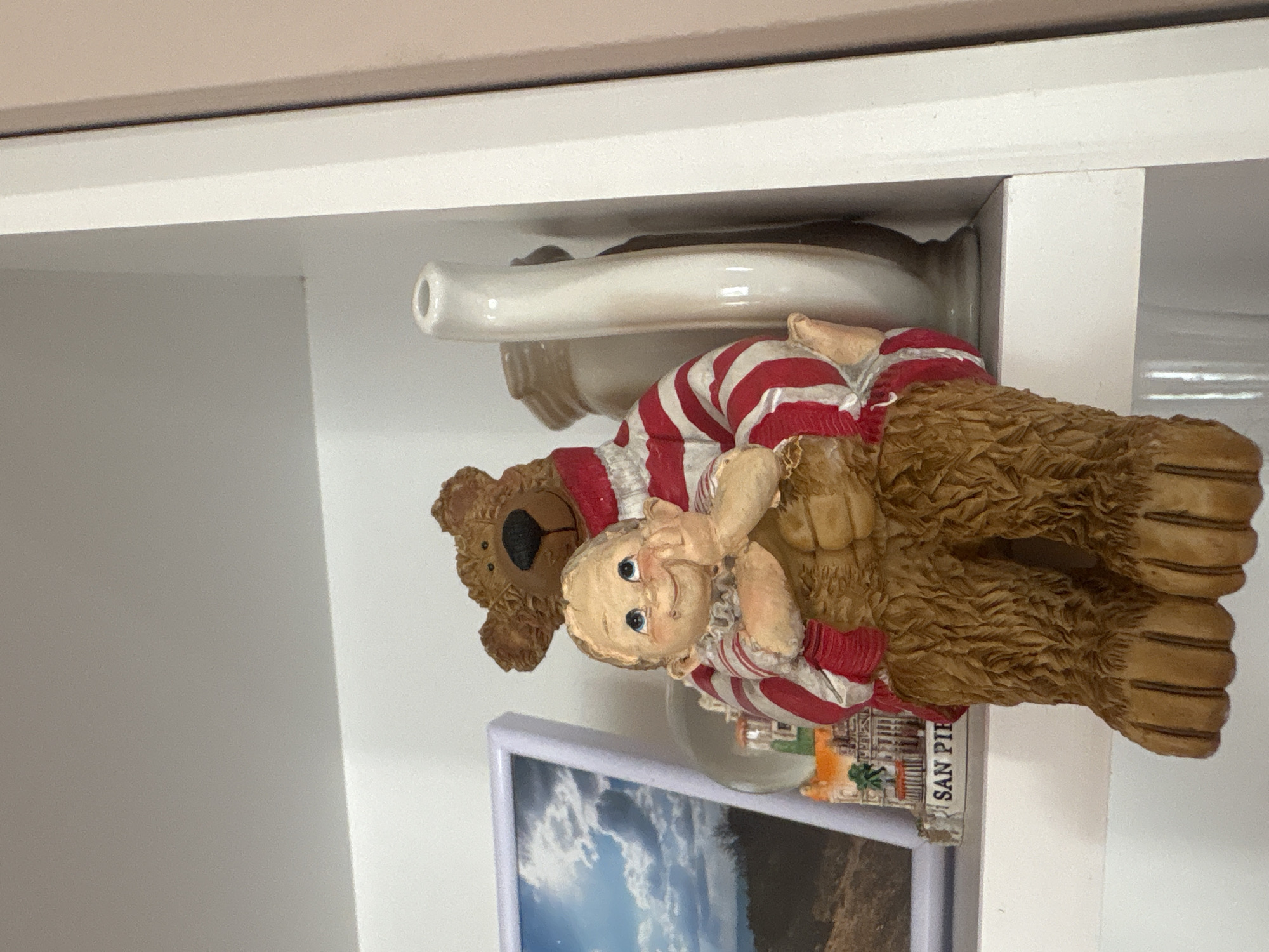 A ceramic figurine of a bear holding a baby is displayed on a white shelf. The bear is wearing a red and white striped sweater, and the baby is also dressed in similar attire. Behind the figurine, there is a white teapot and a framed picture. A small building with the text "SAN PIE" is visible in front of the figurine. The scene appears to be indoors, possibly in a home or shop, but the location in Strelcha, Bulgaria, is not evident from the image itself.