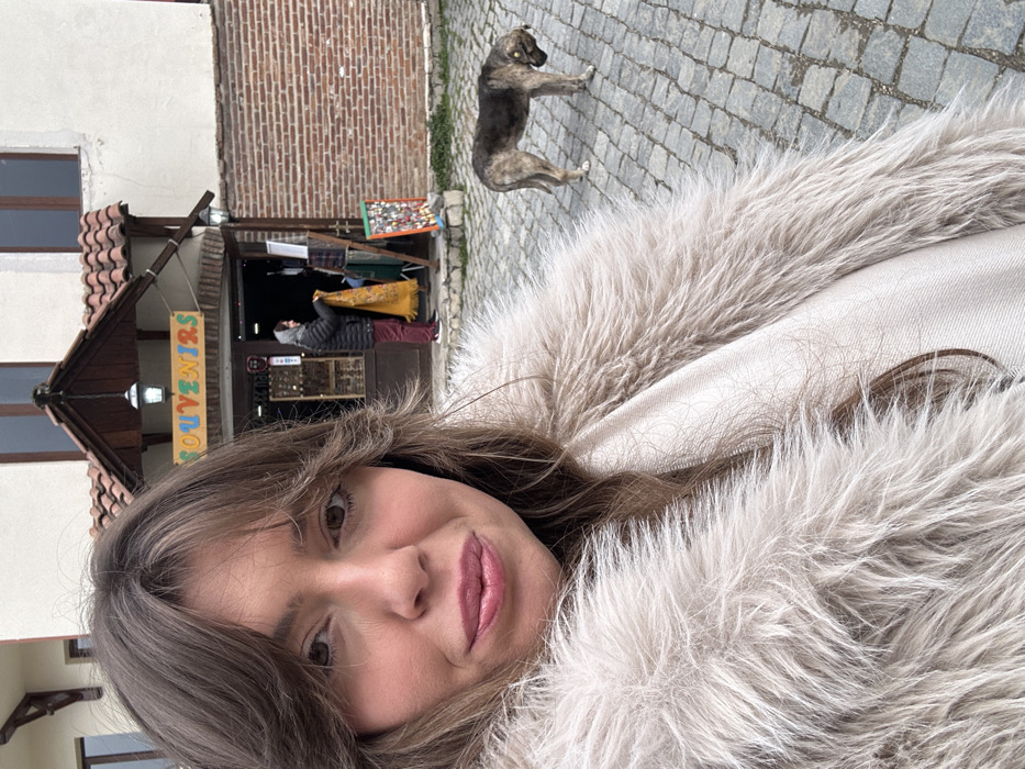 The image depicts a female subject in the foreground taking a selfie in Sighnaghi, Georgia. She is visible from the chest up, with light brown hair and bangs, light complexion, and light pink lipstick. She wears a shaggy, light beige or off-white faux fur coat over a lighter-colored garment. Her expression is a subtle smile as she looks directly at the camera.
In the midground, a "SOUVENIRS" shop is visible behind the subject. The shop entrance is framed in dark wood, with a colorful sign reading "SOUVENIRS" hanging above it. The building features a white plastered upper facade, a red-tiled roof, and two dark wall-mounted lanterns flanking the entrance. Inside the shop doorway, a person wearing a dark jacket and red trousers is observed holding up a yellow patterned textile item. To the right of the shop entrance, an outdoor display stand holds multiple small, colorful items.
To the right of the shop, a medium-sized dog stands on all four legs on the cobblestone pavement. The dog has multi-toned dark and light grey-brown fur and a yellow ear tag visible on its right ear. Its head is lowered towards the ground. The entire visible ground surface is paved with grey cobblestones. The lighting is consistent with daylight, possibly overcast.