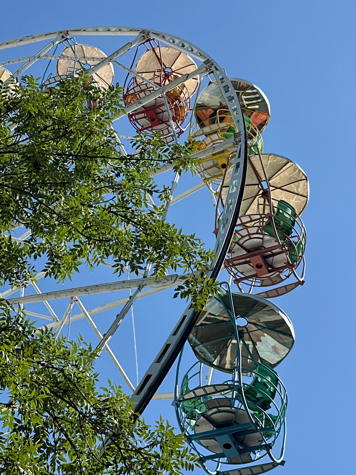 A Ferris wheel, partially obscured by lush green tree branches, stands against a clear blue sky. The Ferris wheel is constructed of white metal beams forming a large circular structure. Attached to the wheel are various gondolas, each with a unique color scheme. Some gondolas have red and tan circular roofs with spokes, while others are enclosed in teal cages with green seating. The gondolas appear to be empty. The image is taken from a low angle, looking up at the Ferris wheel. The setting is an outdoor amusement area in Gyumri, Armenia.