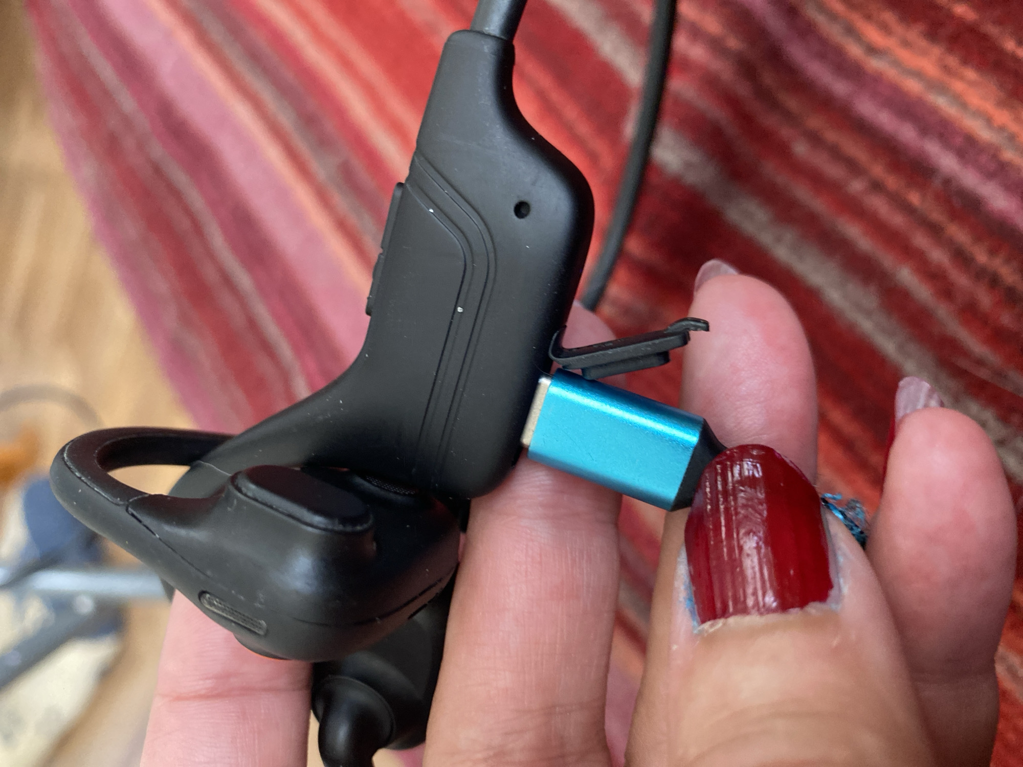 A person is holding a black wireless earbud with a blue charging connector. The earbud is designed to loop over the ear for a secure fit. The charging connector appears to be a USB-C type and is plugged into a small port on the earbud. The person's fingers, with red nail polish, are visible, holding the earbud and connector. In the background, a striped fabric, possibly a blanket or upholstery, with shades of red, pink, and orange, is visible. The lighting suggests it is daytime, possibly indoors, given the close-up nature of the shot and the lack of strong shadows. There is no visible text in the image. The setting appears to be domestic or personal, with the focus entirely on the earbud and the act of connecting it. The image conveys a sense of everyday activity, likely related to charging the device.