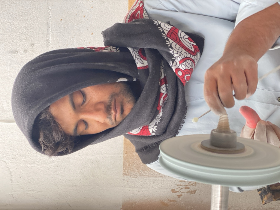 The image depicts an adult male engaged in a detailed manual task within what appears to be a workshop in Kandahar, Afghanistan. The subject has dark hair and a beard, and wears a light blue long-sleeved shirt under a black headscarf with red and white patterns. His eyes are closed, suggesting intense concentration. He is operating a motorized polishing station; a rapidly rotating light-grey wheel is visible on the left. His left hand, protected by a red thimble-like covering on the thumb, stabilizes a light-colored, conical workpiece mounted on a spindle. His right hand precisely positions a slender tool, featuring a small, spherical light-colored tip, against the workpiece. Fine dust or particulate matter is observable on the subject's hands and the immediate work area. The background consists of a plain, light-colored wall with a horizontal brown band.