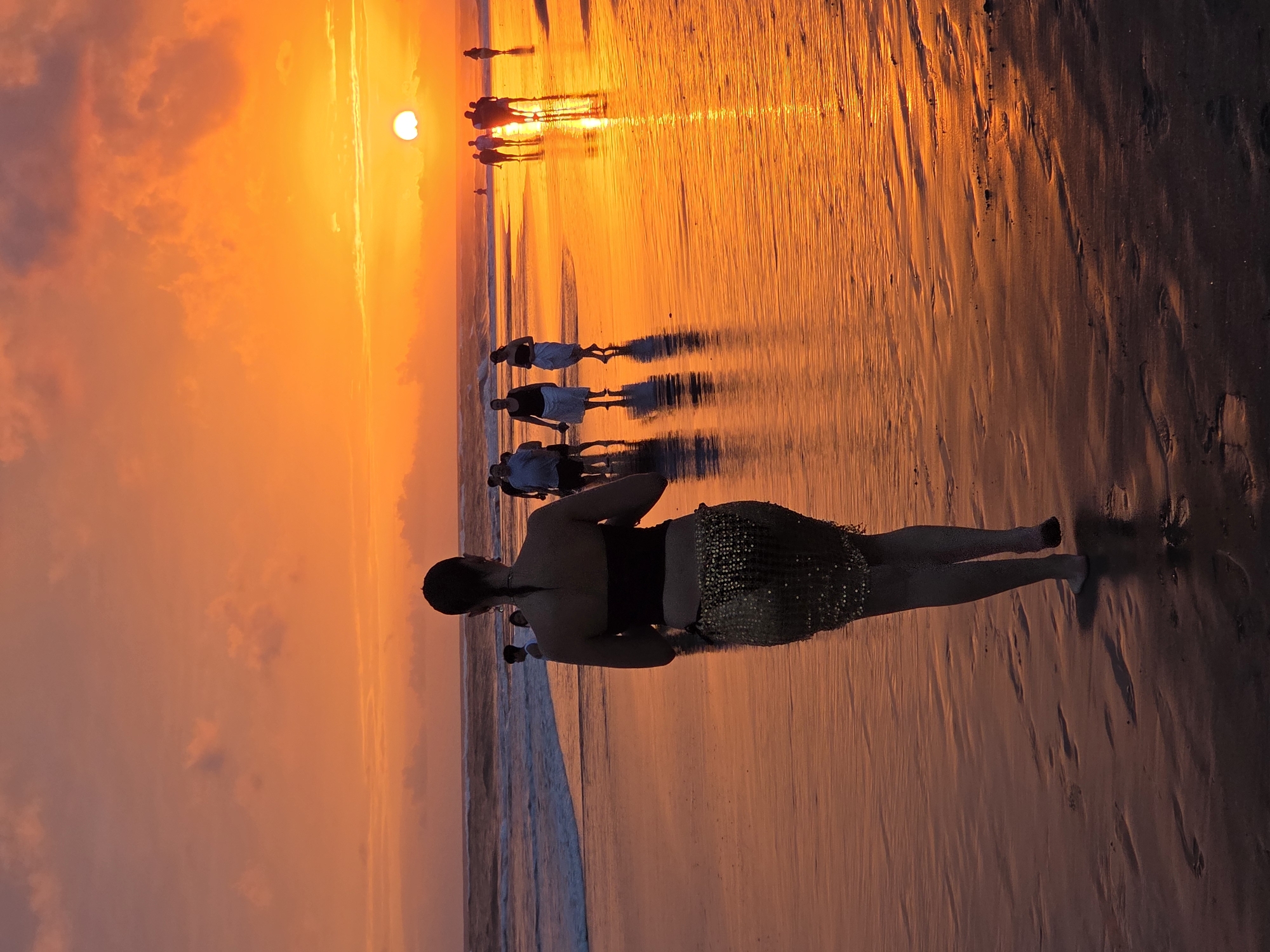 At sunset on the beach in North Kuta, Indonesia, a woman with dark hair pulled back walks away from the camera. She wears a black strapless top and a shimmering gold skirt. The wet sand reflects the vibrant orange sky and the sun, which is setting over the ocean. Several other people are visible in the background, walking along the shoreline, their figures silhouetted against the bright sky and water. Waves gently break on the beach. The scene is bathed in the warm, golden light of dusk.