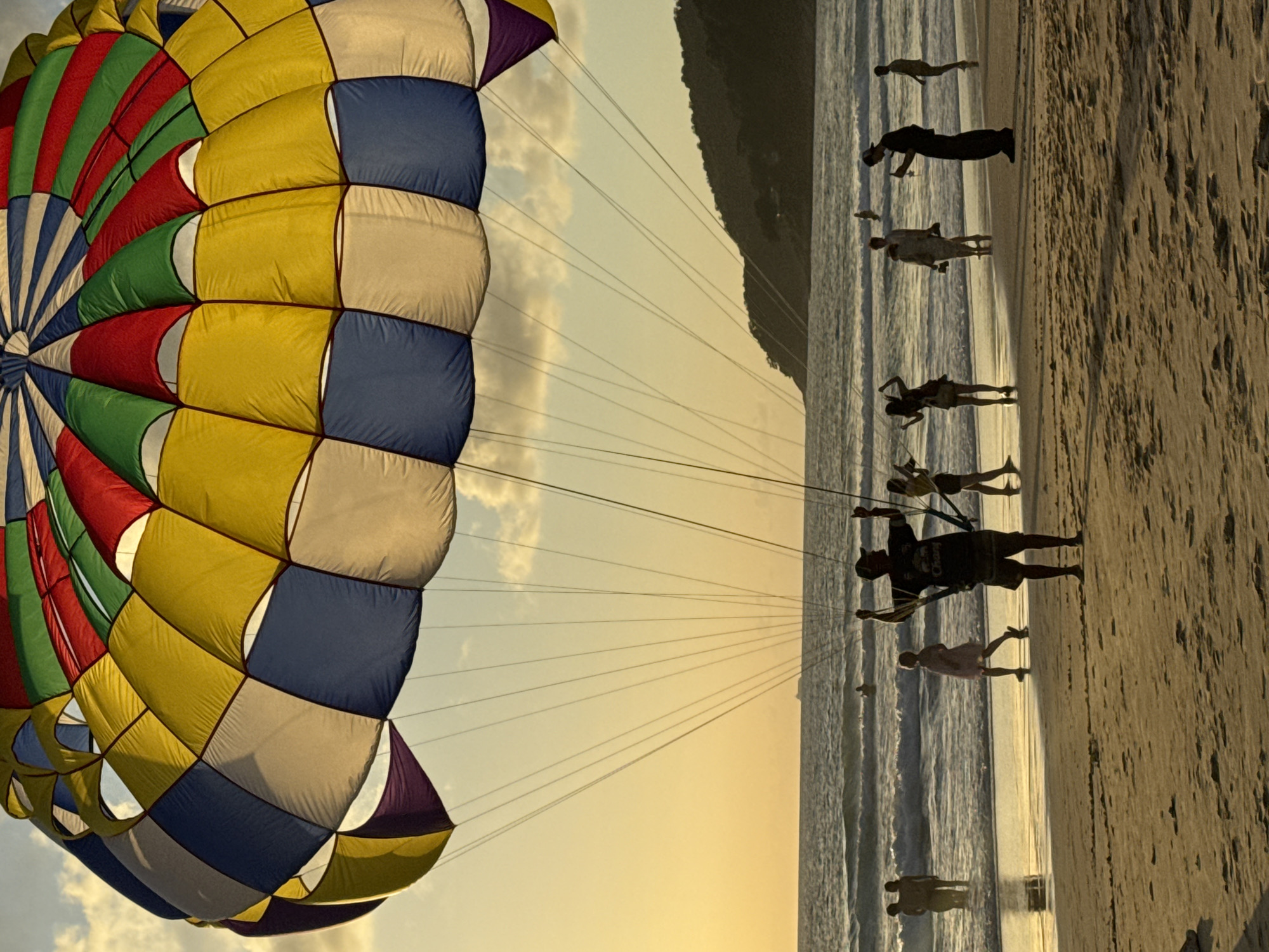 A wide-angle shot captures a colorful parasail against an overcast sunset sky over Karon Beach, Thailand. The parasail, with its alternating yellow, white, blue, red, and green panels, dominates the upper portion of the frame. Below, a shoreline is visible, with gentle waves rolling onto the sandy beach. Silhouetted figures dot the sand and water. In the foreground, a man stands on the beach, facing the water, with his arms raised. To his left, a young girl in a pink dress walks towards the water. To the right of the man, several other figures are visible, including a woman in a yellow top and shorts, and a man in dark clothing holding a phone. In the distance, several more people are seen in the shallow water, and a faint outline of a ship is visible on the horizon. The overall atmosphere is one of leisure and recreation.
