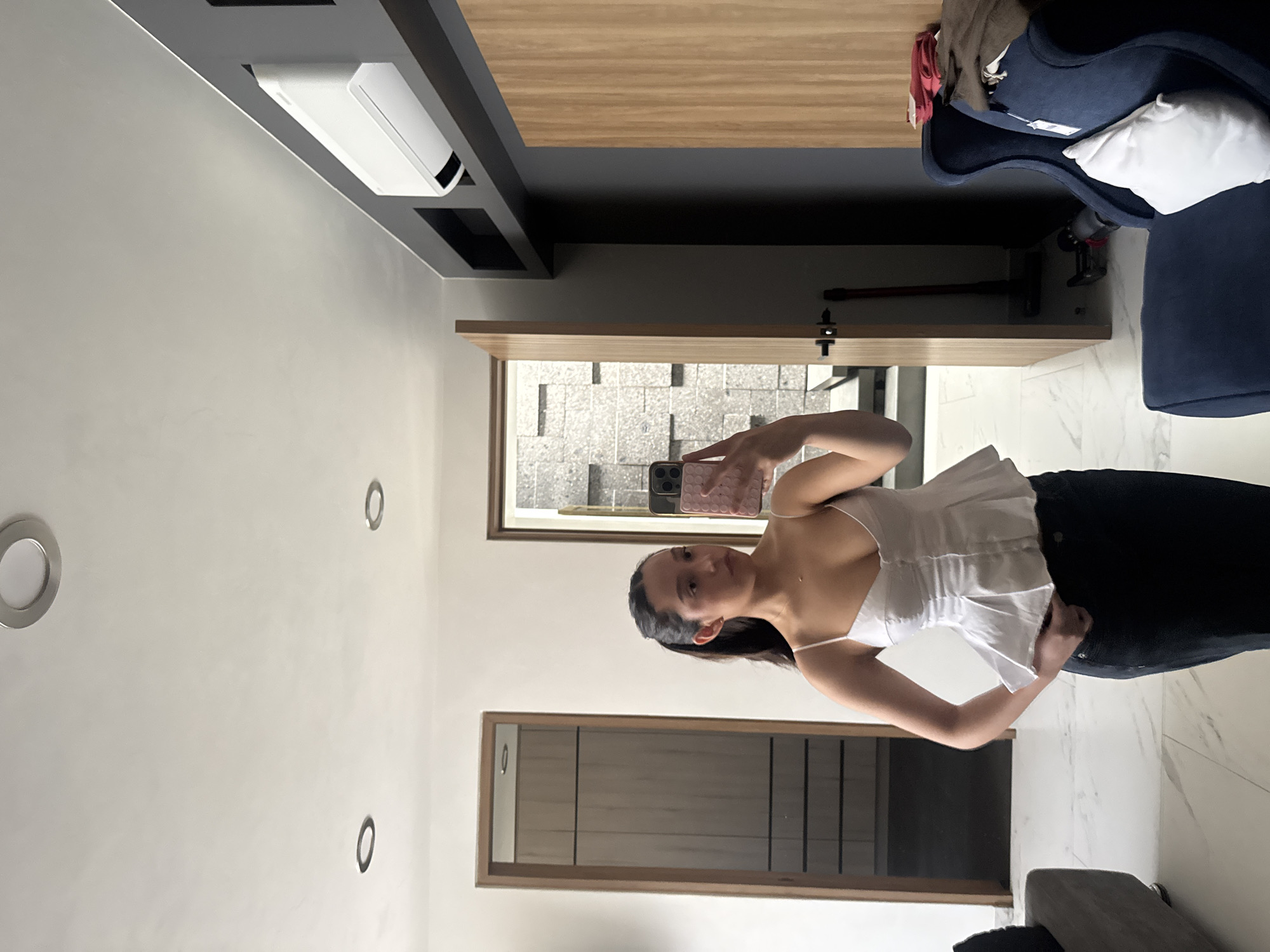 The image displays a young woman taking a mirror selfie in a modern indoor setting. The woman stands upright, holding a smartphone with a pink, textured case in her left hand, which is raised to take the photo. She wears a white, sleeveless top with a ruffled hem and dark-colored pants. Her dark hair is pulled back, and she looks directly into the camera with a neutral expression.
The room features light gray walls and a light-colored tiled floor, possibly marble. Several circular recessed lights are visible on the ceiling. High on the wall, a white rectangular air conditioning unit is installed within a dark gray shelving or structural element, which also supports a wooden panel extending across the upper part of the room.
To the right, a dark blue armchair or sofa is visible, topped with a white pillow and some folded clothes. Behind the armchair, a black stick vacuum cleaner leans against the wall. Behind the woman, a large wooden-framed opening reveals an outdoor area with patterned gray paving, suggesting a patio or courtyard. Another wooden-framed doorway or opening is visible further to the left, showing a glimpse of an adjacent room.
The setting appears to be a contemporary living space, likely during the daytime, given the natural light visible from outside. No visible text is legible in the image. The modern decor and outdoor paving are consistent with a residence in a location like Puebla, Mexico.