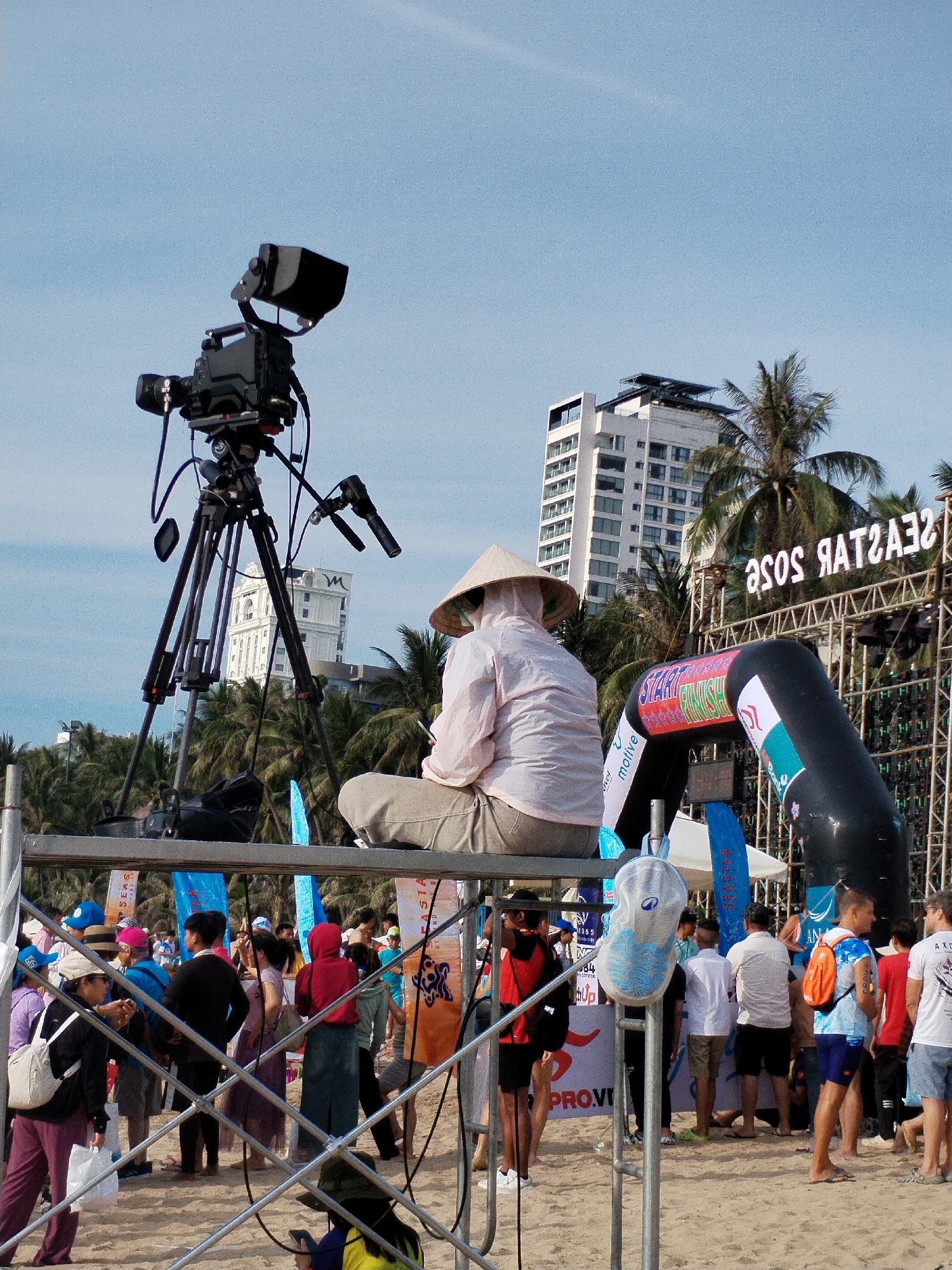 This image depicts a scene on a beach in Nha Trang, Vietnam, likely during an outdoor event. In the foreground, a large professional video camera on a tripod is positioned prominently. To the right of the camera, a person wearing a conical hat and a light-colored shirt is seated, facing away from the viewer. Behind them, an inflatable archway with "START FINISH" printed on it marks a race or event entrance. Further in the background, a large stage structure with illuminated signage spelling "SEASTAR SOSE" is visible, along with palm trees and modern buildings. A crowd of people, some in athletic wear, are gathered on the sandy beach, suggesting a sporting event or festival is underway. The sky is clear and blue.