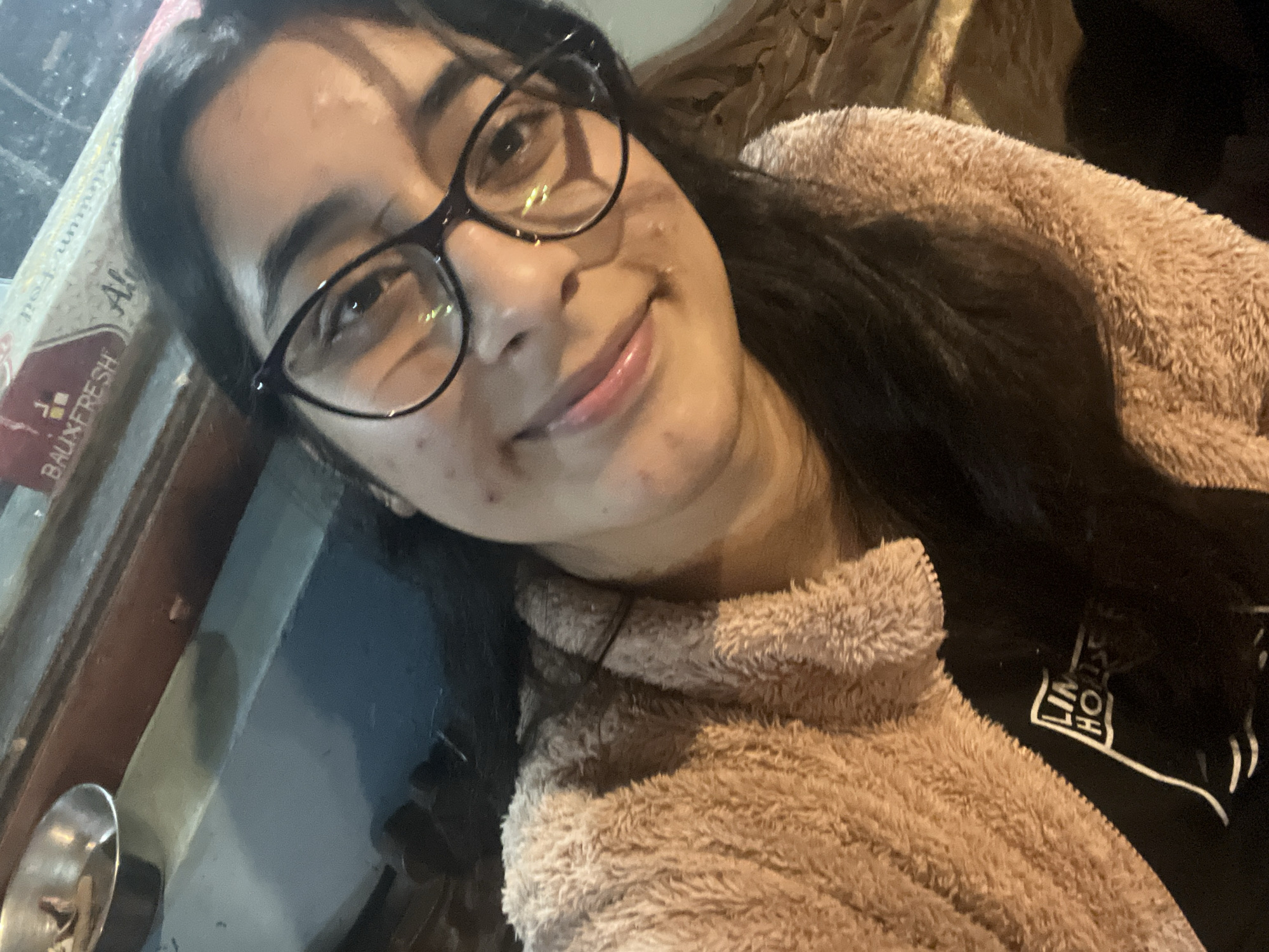 A young woman with dark hair, wearing glasses and a fuzzy pink jacket, takes a selfie. She is smiling and appears to be indoors, possibly in a restaurant or cafe, given the presence of a metal bowl with items in it and a roll of aluminum foil in the background. The lighting is warm and suggestive of an evening setting. Her black t-shirt underneath the jacket has a white logo that reads "LIN HO SE".