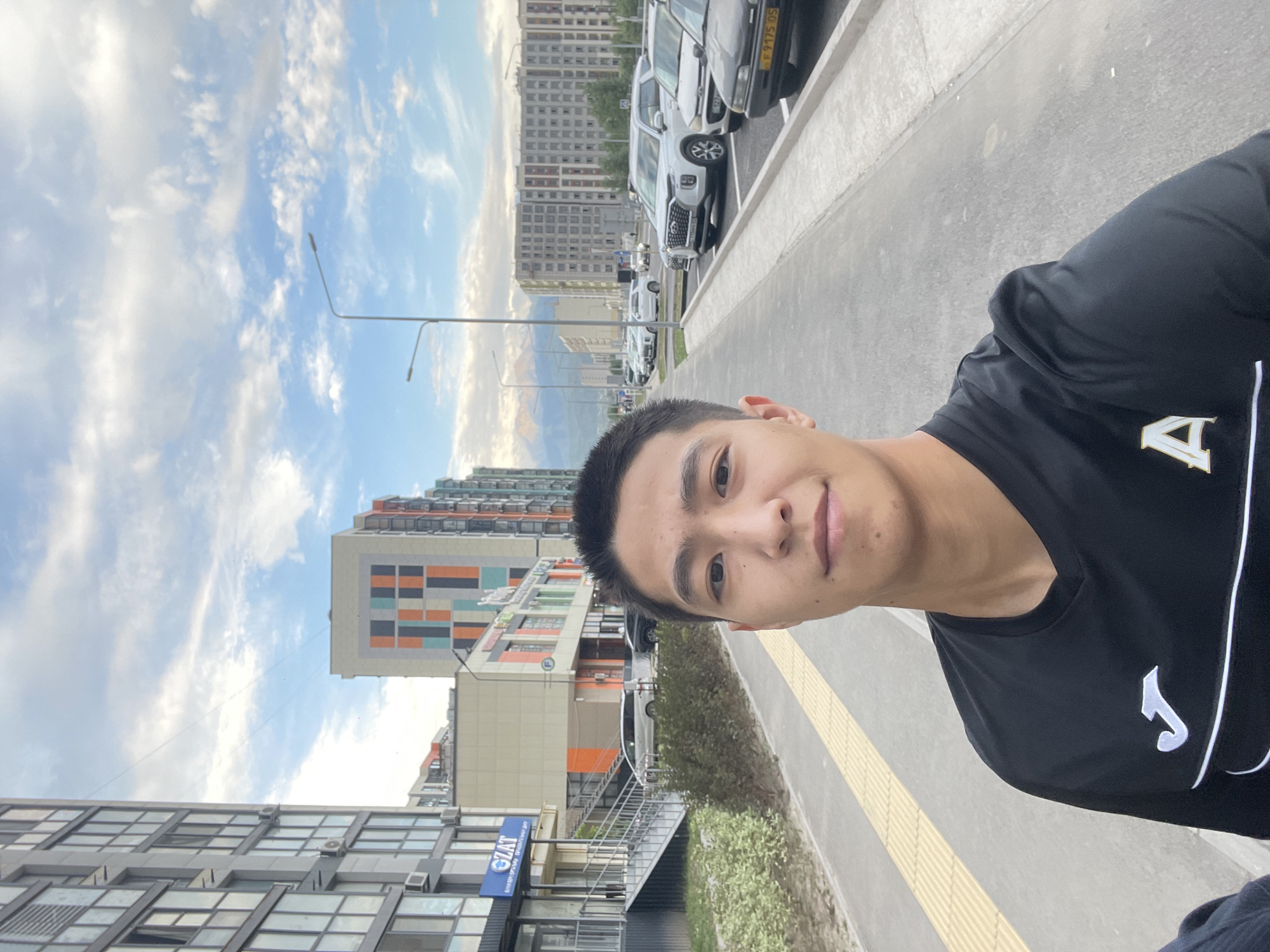 A young man with short black hair and a light complexion is taking a selfie outdoors. He is wearing a black long-sleeved shirt with white and gold logos on it. The background suggests an urban environment with modern, multi-story buildings. To the left, a tall building with many windows is visible, and to the right, a street lined with parked cars and other buildings stretches into the distance. In the far background, mountains are faintly visible under a cloudy sky. The sidewalk in front of the man appears to be a paved pathway with tactile paving for visually impaired pedestrians. The overall scene depicts a contemporary residential or commercial area in Almaty, Kazakhstan.