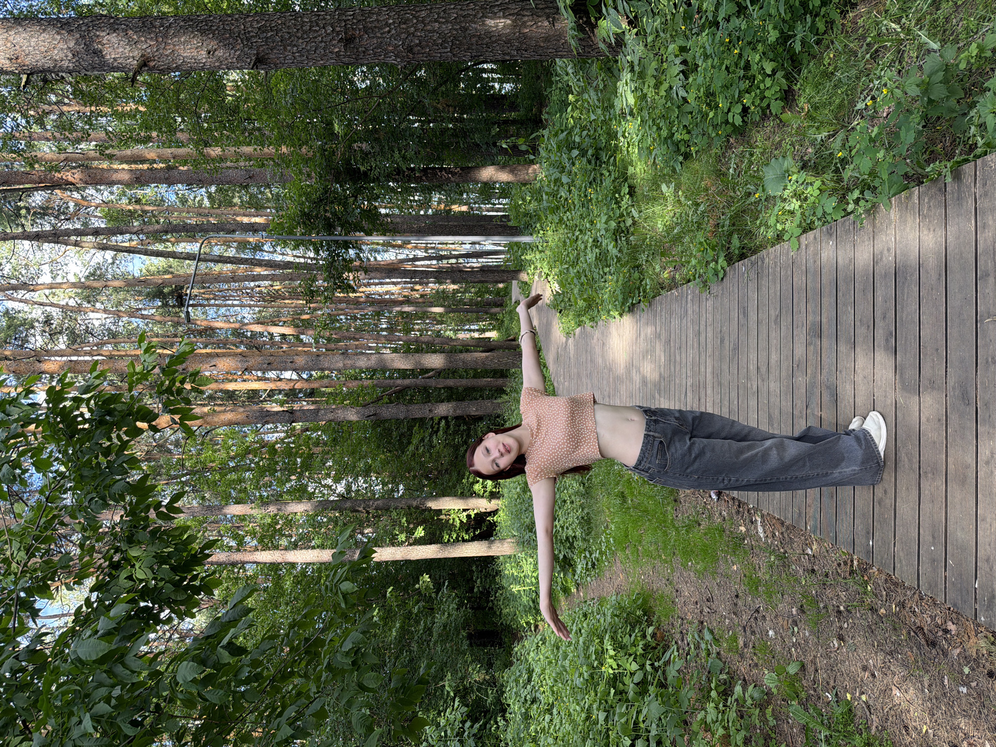 A young woman with long, dark hair and fair skin is captured in a full-body shot, lying on her back on a wooden boardwalk. She is wearing a light pink crop top with small white polka dots and dark grey wide-leg jeans. Her arms are outstretched, one hand reaching upwards and the other reaching downwards, as if balancing or in a relaxed pose. She is looking directly at the camera with a neutral expression.
The setting is a sun-dappled forest with tall pine trees lining the boardwalk. The boardwalk itself is made of weathered wooden planks and curves gently to the right, disappearing into the greenery. Lush green foliage and grass flank the boardwalk, with some small yellow flowers visible among the plants. The lighting suggests it is daytime, likely late morning or early afternoon, with sunlight filtering through the trees and casting shadows.
The scene appears to be in a natural park or forest area, possibly a recreational trail, given the constructed boardwalk. The overall mood is peaceful and serene, with the woman appearing comfortable and at ease in her natural surroundings. No visible text is present in the image. The location is Chaikovsky, Russia.