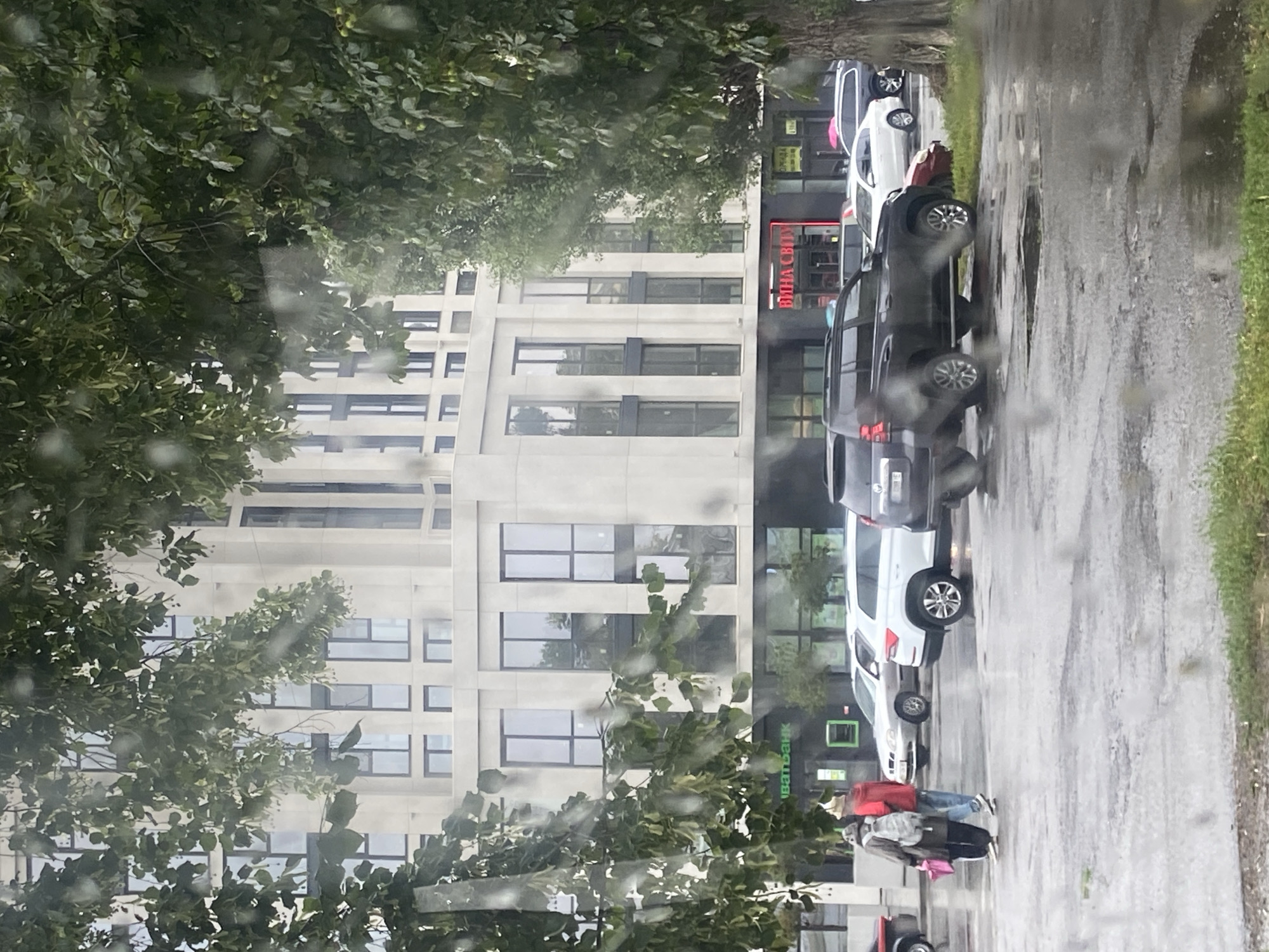 The image depicts a street scene in Nyzhni Mlyny, Ukraine, on a rainy day, as evidenced by water droplets on the lens and wet pavement. In the foreground, a tree with green leaves partially obscures the view. Behind the tree, a modern, light-colored building with numerous windows stands tall. At the base of the building, there are storefronts. One storefront displays green lettering that reads "ІВАТБАНК," indicating a bank. Another storefront has red neon lettering spelling "ВИНА СВІТУ," suggesting a wine shop.
In front of the building, a parking lot is visible with several cars. A dark-colored SUV is prominently parked. Two individuals, one wearing a red jacket and a hat, and another in a beige jacket and a backpack, are walking away from the viewer towards the building, carrying a pink bag. The ground is wet and reflective, with visible puddles. The overall atmosphere is subdued due to the overcast weather.