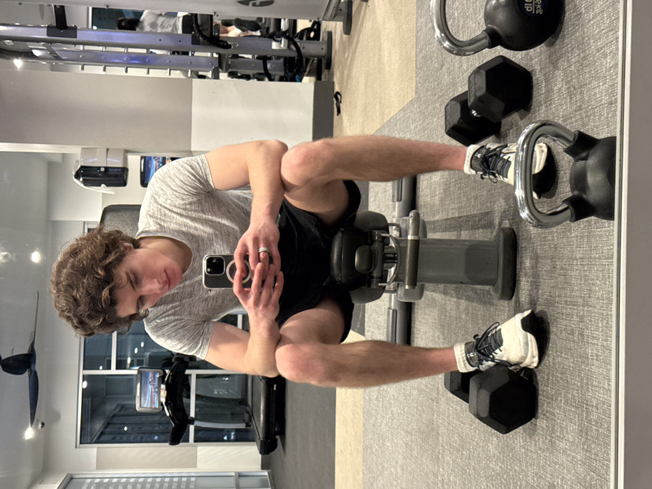 The image captures an indoor gym facility, identified as being in Los Angeles, United States of America, though specific visual identifiers for the city are not discernible from the visible cityscape.
A young adult male, characterized by medium-length curly brown hair, fair complexion, and a lean physique, is seated centrally on a grey adjustable exercise bench. He is attired in a light grey short-sleeved t-shirt, dark athletic shorts, white athletic socks, and white and black athletic shoes. A silver-colored ring is present on his left ring finger, and a single earbud is visible in his right ear. Both hands hold a dark-colored smartphone, which displays a circular magnetic attachment on its rear, and he is looking intently at the screen.
The floor is covered with a patterned grey carpet tiling. In the foreground, a black kettlebell with a silver handle is partially visible, displaying "16LB" text. Two black hexagonal dumbbells are positioned on the floor, one on each side of the male's feet.
The background features large windows offering a view of an urban landscape illuminated by city lights, indicating evening or night conditions. To the left, a black treadmill with an active digital display screen is visible. A dark-bladed ceiling fan is mounted overhead. Towards the right, a white wall-mounted dispenser is present, and gym equipment, including a grey multi-station weight machine, is visible, partially as a reflection. A reflection of another individual, seen from behind wearing a light-colored top and dark bottoms, is also visible on the right, suggesting additional gym users. The facility is well-lit by artificial lighting.