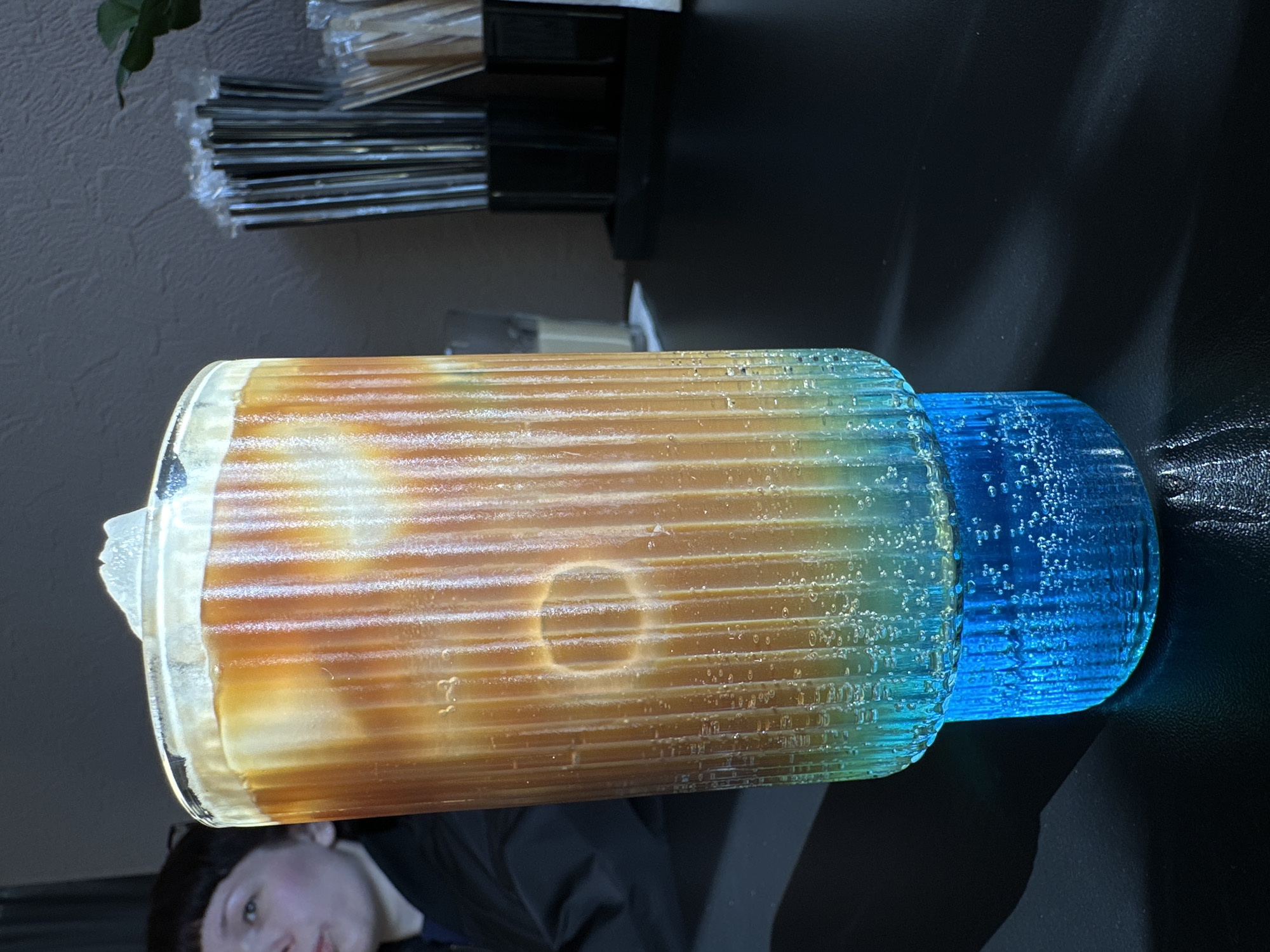 A horizontally oriented close-up shows a clear ribbed glass. The top part of the glass is filled with a tan liquid, and the bottom part is filled with a blue liquid with visible air bubbles. The glass sits on a black surface. Behind the glass, there is a white wall with a green plant in the upper left corner and a container holding black-and-silver straws. A person's face is partially visible in the lower left corner. The location is not discernable from the image.