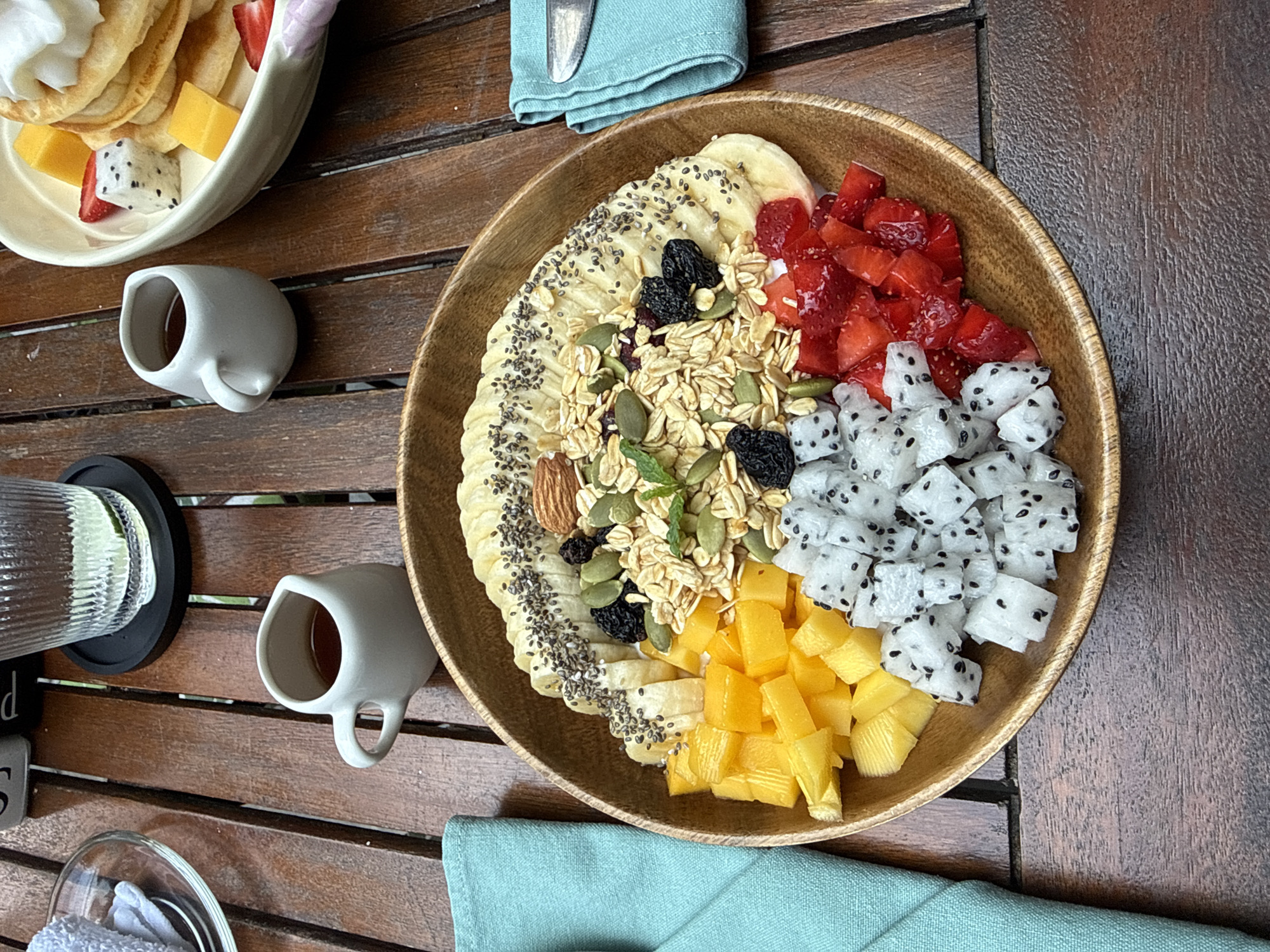 The image captures a breakfast scene, likely at a restaurant or cafe in Đà Nẵng, Vietnam. The scene is set on a dark wooden table, typical of outdoor dining furniture.
A large wooden bowl dominates the center of the image. It contains a variety of breakfast items, arranged segmentally. From the top left, there are sliced bananas topped with chia seeds, followed by a mixture of granola, pumpkin seeds, almonds, and dried cranberries. Clockwise, there are sections of diced strawberries, cubed dragon fruit, and diced mango.
In the upper left corner, a shallow white bowl contains pancakes, topped with whipped cream and pieces of mango, strawberries, and dragon fruit.
There are two small white ceramic cups with handles, likely containing coffee or tea, placed on the table.
Adjacent to one of the cups is a tall, ridged glass pitcher, possibly containing water.
A folded light blue or green napkin sits near the wooden bowl, and another napkin lies partially under the bowl. Cutlery is visible resting on one of the napkins.
The background is blurred, suggesting a depth of field effect, but the setting appears to be outdoors. The lighting suggests a bright, sunny day, typical of Đà Nẵng's climate. There are some letters written in grey on a grey slab on the bottom left part of the image, which are unreadable.