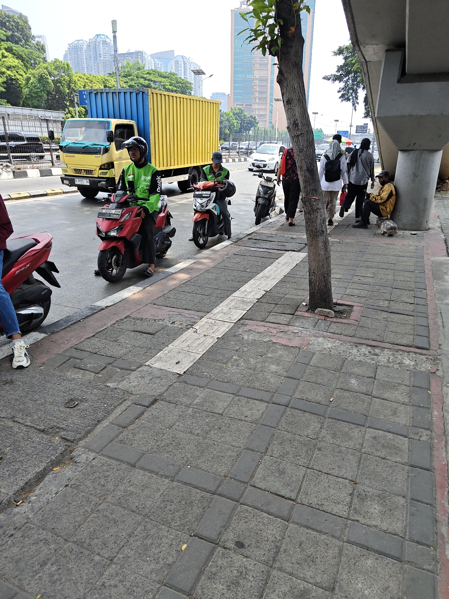 In West Jakarta, Indonesia, a street scene unfolds under an overcast sky. A yellow truck with a blue container travels on the road, accompanied by several motorbikes. Two individuals on motorbikes, wearing green jackets and helmets, are positioned on the road. One is on a red motorbike, and the other on an orange motorbike. In the background, buildings of varying heights rise against the horizon, with lush green trees framing the urban landscape. A concrete pedestrian walkway runs alongside the road, marked by gray paving stones and a red border. Several people walk along the sidewalk. A man in a yellow jacket sits by a large concrete pillar, leaning against it. Another person is seated on the ground near the pillar. The foreground shows a close-up of the pedestrian walkway with its textured paving stones.