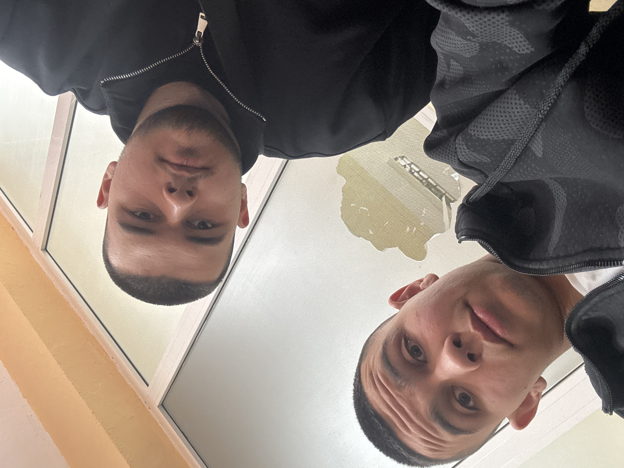 Two men are depicted from a low-angle perspective, suggesting the camera is positioned below them, looking up. The man on the left wears a black jacket with a visible zipper. He has short dark hair and a light beard. The man on the right wears a jacket with a camouflage pattern. His hair is also short and dark. In the background, a building and windows are visible, reflecting light. The location is in Varna, Bulgaria based on the architecture of the building.