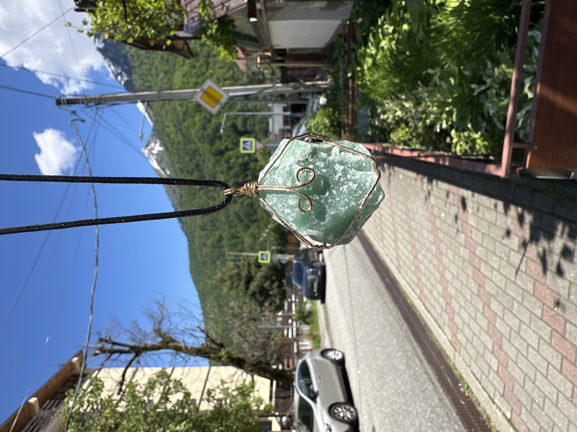 A rough, light green crystal pendant, intricately wrapped in copper wire and suspended by a black braided cord, hangs prominently in the left foreground of the image. The scene unfolds on a bright, sunny day in Krasnaya Polyana, Russia.
The background reveals a street with an asphalt road and a patterned brick sidewalk on the right. Lush green vegetation, including trees and bushes, lines the side of the road, behind which buildings are partially visible. Several cars are parked along the street, with a silver car closer to the viewer and a darker one further back.
In the mid-ground, a tall utility pole with numerous power lines stands, alongside two road signs: a yellow diamond-shaped sign, typically indicating a main or priority road, and a blue square sign with a white pedestrian crossing symbol. Beyond the immediate street, majestic mountains, heavily forested with green trees and showing patches of snow near their peaks, dominate the distant horizon under a clear blue sky adorned with scattered white clouds.
The bright daylight suggests it is midday, with strong shadows visible on the sidewalk. There is no explicit human activity depicted, focusing instead on the pendant against the scenic mountain town backdrop. No specific text is legible on the signs or other objects.