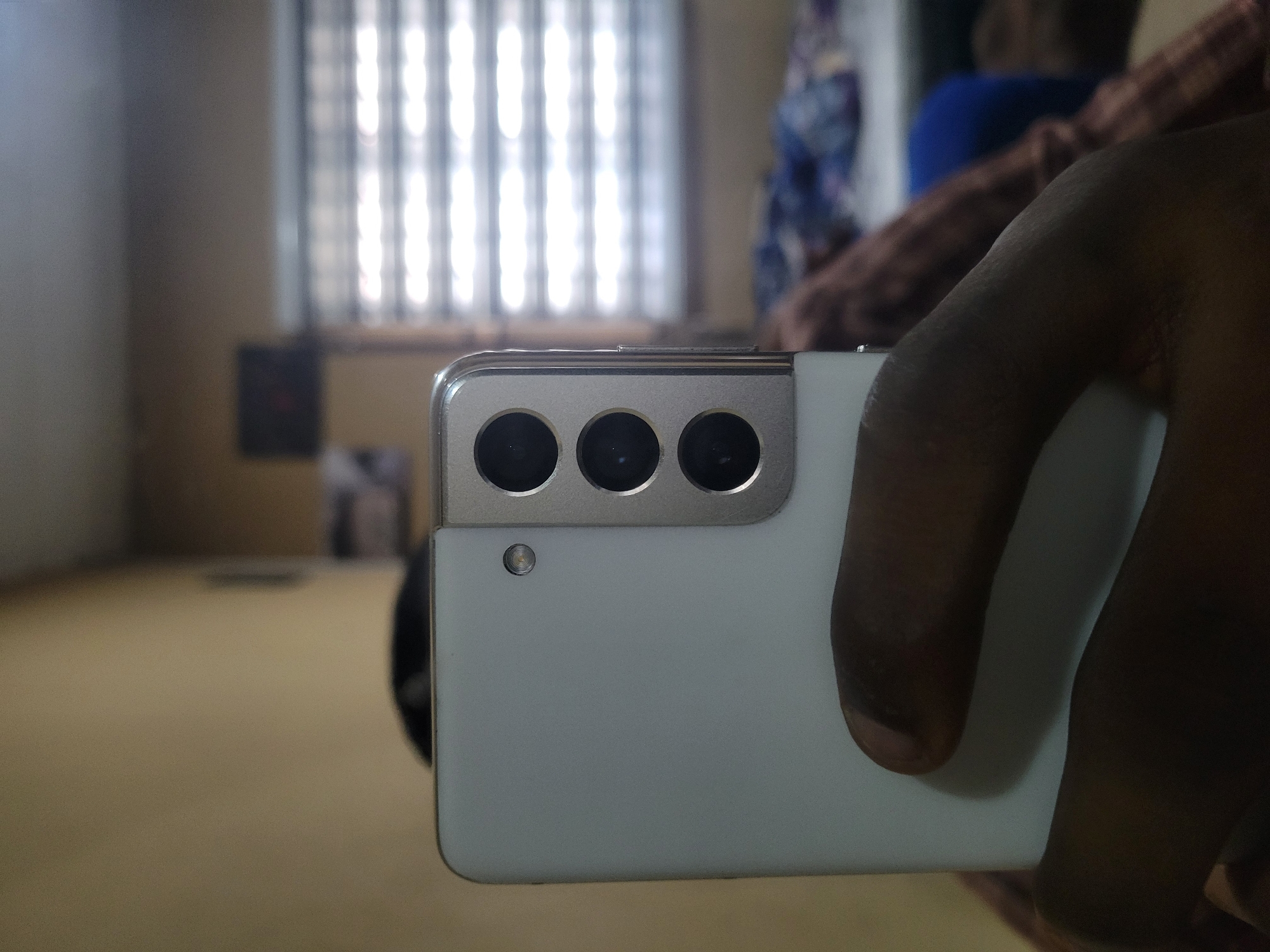 A close-up, eye-level, slightly angled shot shows a person's hand holding a white smartphone. The back of the phone is visible, featuring a rectangular camera module with three circular lenses arranged vertically, and a small circular flash or sensor above the top lens. The phone's casing appears to be made of a smooth, matte material with a metallic rim around the camera module.
The background is out of focus, but a window with horizontal blinds is visible, letting in bright light, suggesting it is daytime. There are some blurred objects in the far background that resemble a bed with blankets. The walls appear to be a neutral, yellowish-beige color. The lighting in the scene is somewhat dim, with a spotlight effect coming from the window. The overall impression is that of an indoor setting, possibly a bedroom or living area, in a location that cannot be determined.