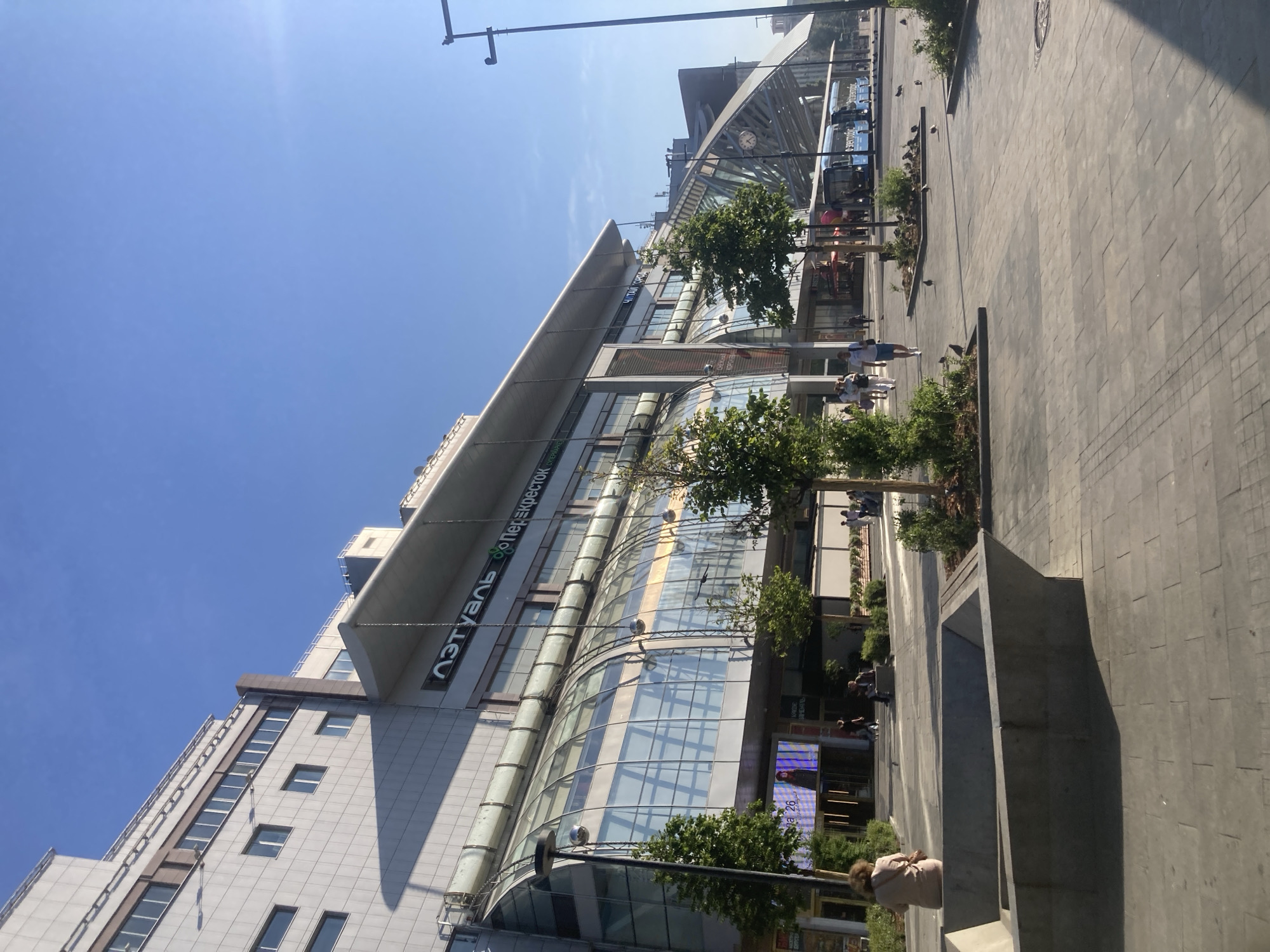 The image depicts a sunny day in an urban environment, likely a shopping district in Moscow, Russia. A large, modern building with a glass-covered entrance and a curved roof dominates the scene. The building displays prominent signage, including "Л'Этуаль" (L'Etoile) and "Перекрёсток" (Perekrestok), indicating it houses a cosmetics store and a supermarket, respectively.
In the foreground, a paved pedestrian area is visible, with trees and modern concrete planters. Several people are seen walking and sitting in this area. Some are browsing shops, while others relax on the benches. Pigeons are scattered across the pavement.
To the right, a bus stop is situated under a glass-covered structure. Two blue buses are parked at the stop, with one displaying the text "Электрический" (Electric). A traffic light pole is also visible. The sky is clear and blue, indicating fair weather. The overall impression is that of a busy, well-maintained public space.