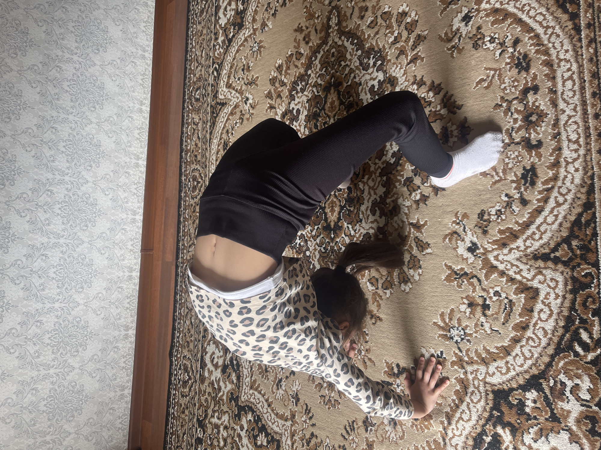A young person, likely a child, is depicted from a top-down perspective performing a backbend or bridge pose on a patterned rug indoors. Their body is arched upwards, with their head facing downwards towards the rug. Dark hair is pulled back into a ponytail. They are wearing black ribbed leggings, a long-sleeved top with a beige, brown, and black leopard print, and white socks. The midriff is exposed, showing the navel.
The activity suggests a moment of exercise, stretching, or playful movement. The rug features an ornate, traditional pattern in shades of beige, brown, and black, covering the floor. To the left, a wall with a light-colored, intricate floral or damask pattern is visible. A dark wooden door frame or trim separates the wall from the rug. The lighting is bright and natural, indicating it is daytime. The scene is set in an indoor environment, specifically within a residence in Poltava, Ukraine. No text is visible in the image.