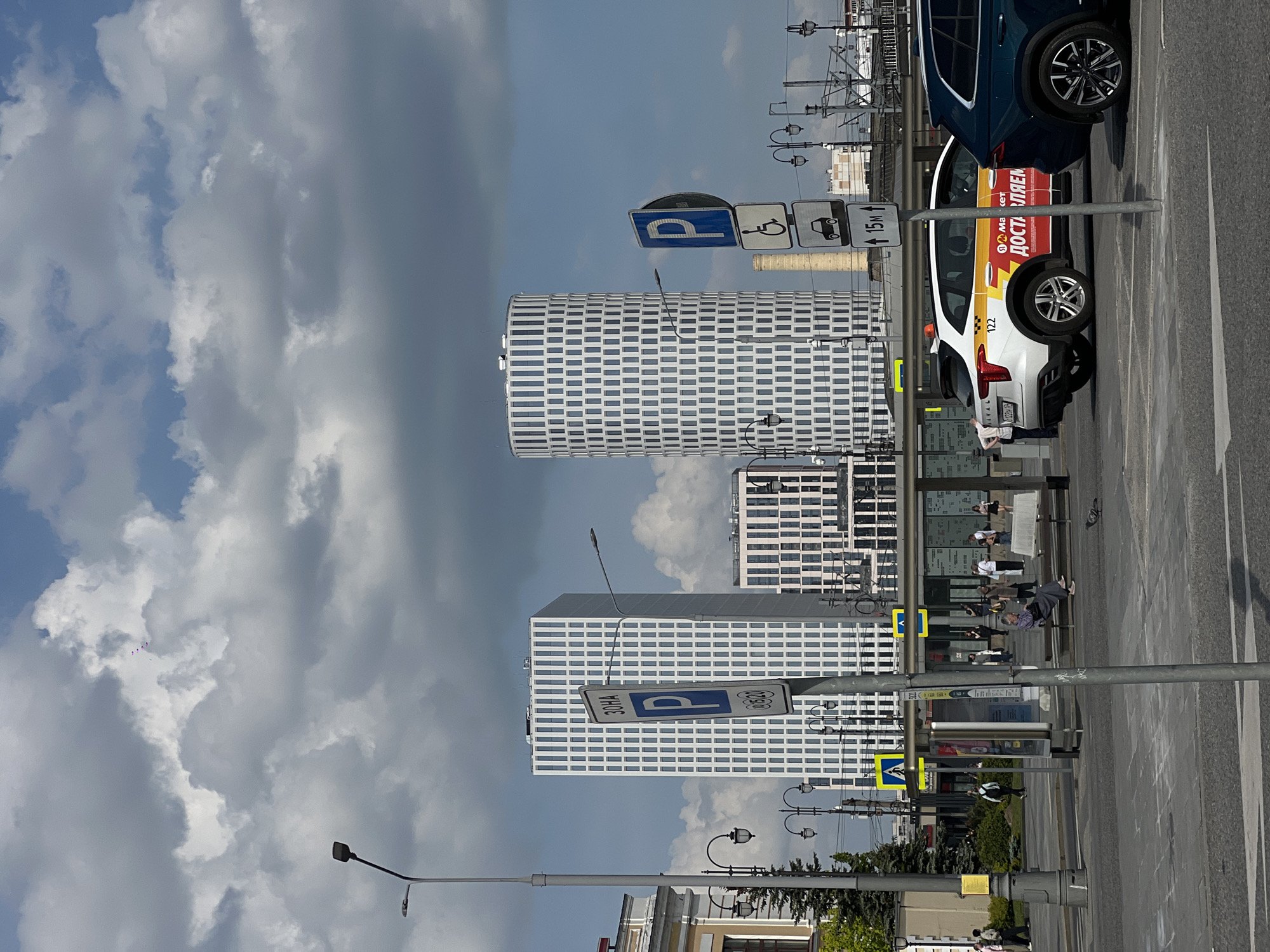 The image shows an urban scene in Moscow, Russia. In the foreground is a bus stop with several people seated. Street lights and parking signs are visible. Behind the bus stop, there are modern, high-rise buildings with a distinctive grid-like facade. Cars are parked adjacent to the curb. The sky is overcast with clouds.