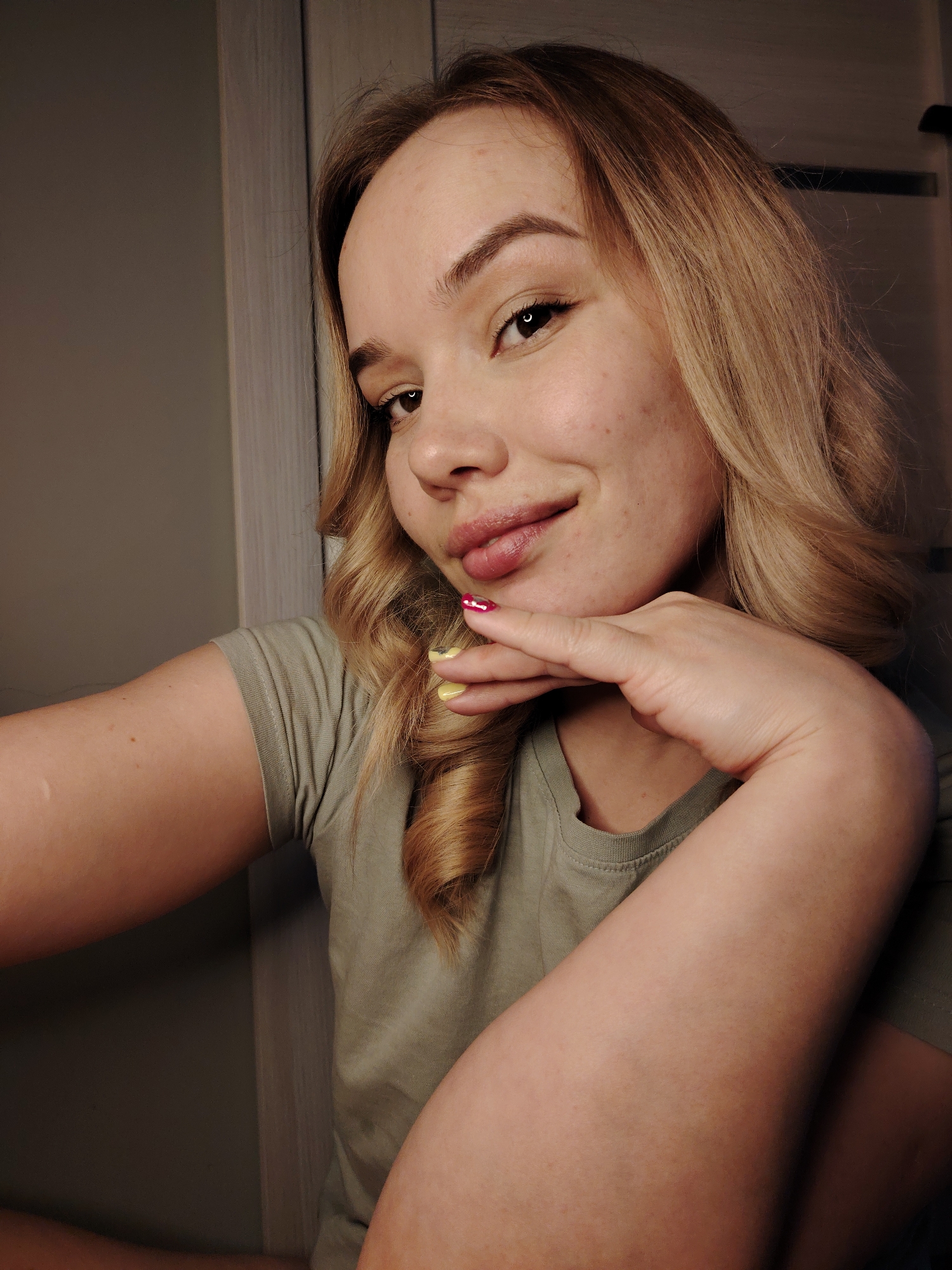 A young woman poses for a selfie in what appears to be an indoor setting. She has fair skin, light brown hair with blonde highlights styled in waves, and is wearing a plain, light green t-shirt. Her left hand is raised, with her chin resting on her fingertips. Her nails are painted with different colors. The background includes a neutral-colored wall and a door with white trim. This image does not provide any information to suggest it was taken in Марковский, Russia.