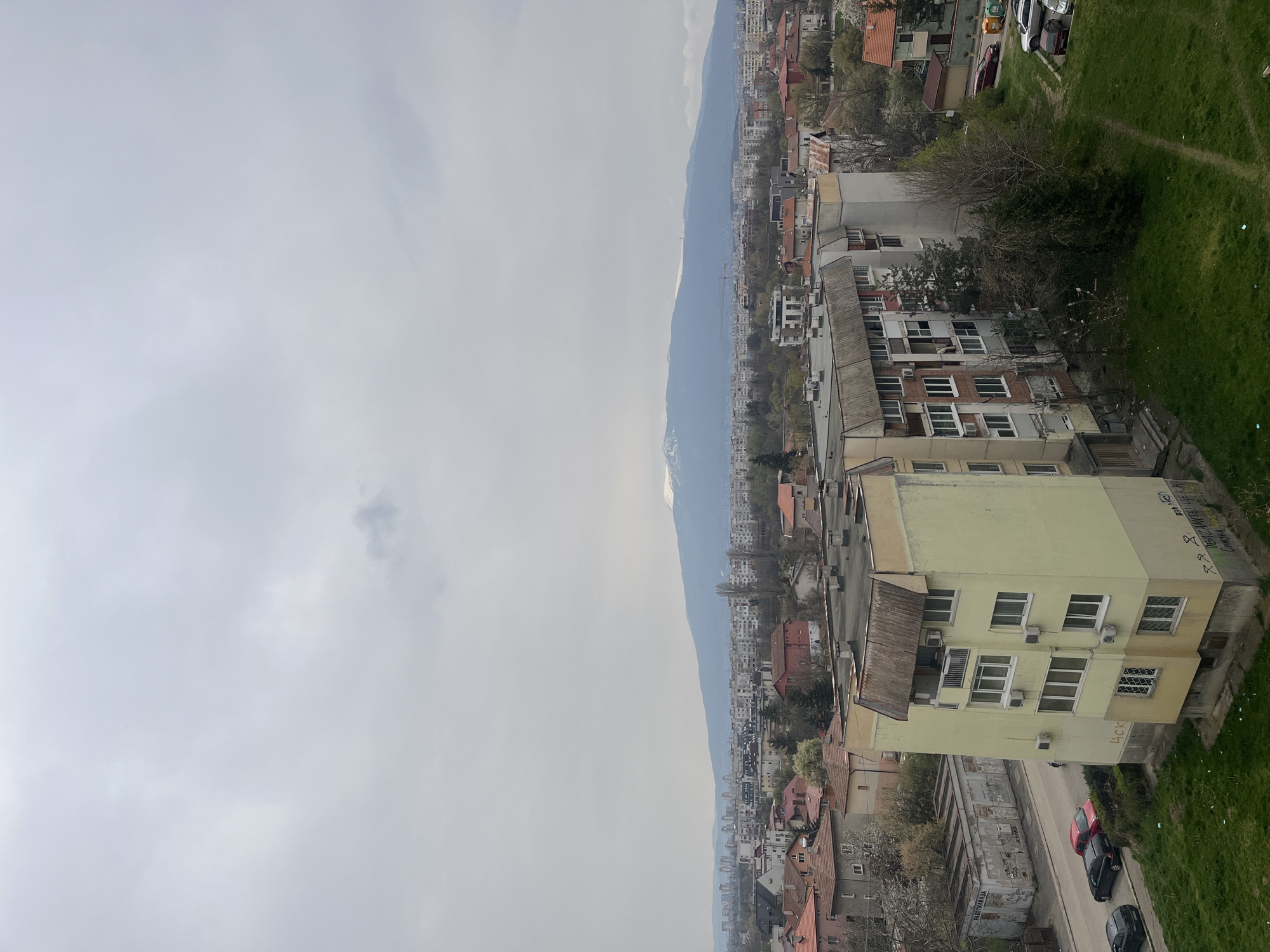 The image shows a view of Sofia, Bulgaria, with a pale overcast sky taking up half the frame. A mountain range, possibly Vitosha, is visible in the distance. The urban landscape is composed of buildings with mixed architectural styles, ranging from residential houses with red-tiled roofs to taller apartment buildings with balconies and air conditioning units. The buildings are situated along a street, where a few parked cars are visible. A patch of green grass borders the edge of the scene, suggesting a park or open space. Some of the buildings have graffiti. There are no visible people.