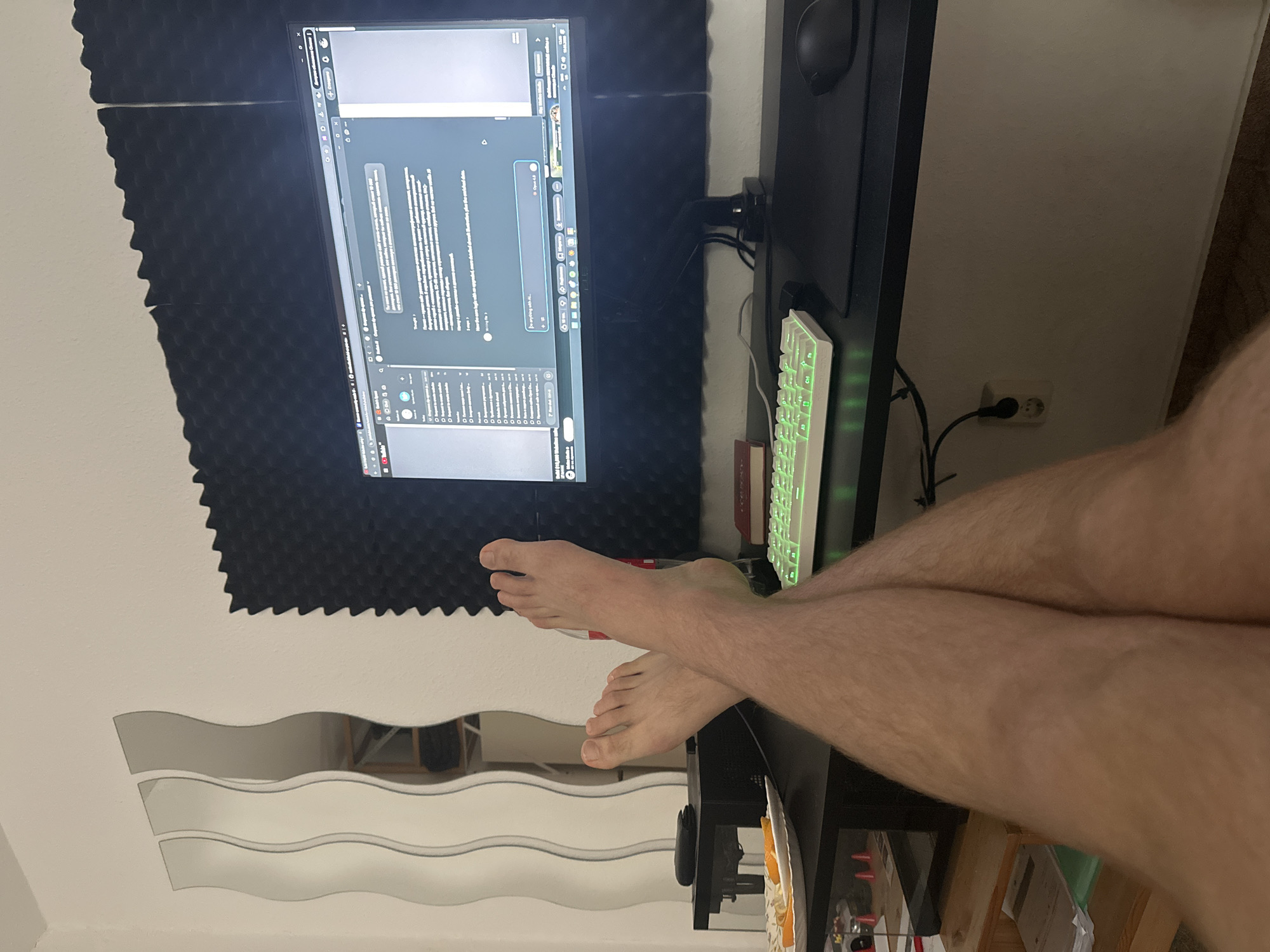 The image depicts a person's legs and feet crossed while sitting at a desk in what appears to be a home office or study. A computer monitor displays text, likely a chat or coding interface, with a green-lit keyboard in front of it. To the left, a plate with orange slices sits on the desk. In the background, a wall is covered with black acoustic foam panels, and a set of three wavy mirrors are mounted on the wall. The overall setting suggests a personal workspace, possibly in Aschersleben, Germany, given the context provided.