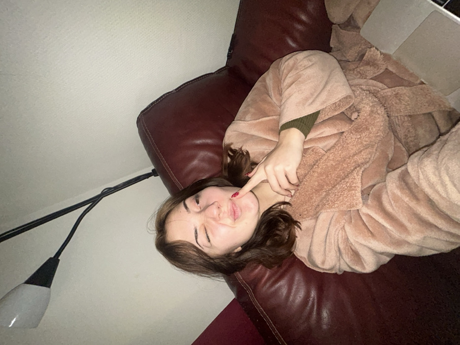 A woman reclines on a maroon leather couch, wearing a fluffy tan robe over a green long-sleeved shirt. She points her finger to her cheek while squinting her eyes and pursing her lips. A floor lamp with a white shade and black base stands to the left. A laptop rests on the couch near her. The background consists of a plain white wall.