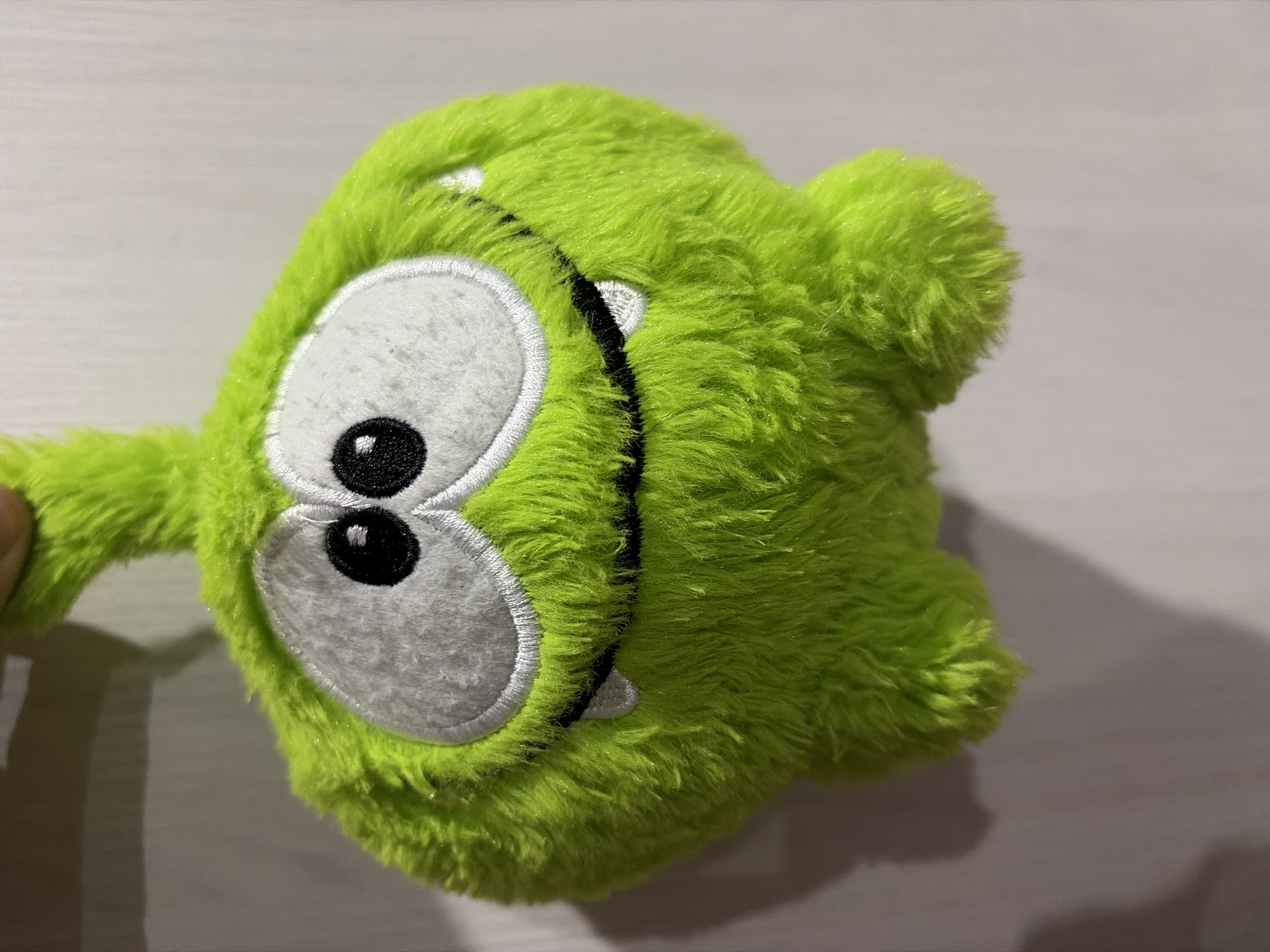 A close-up, slightly above eye-level studio shot features a bright green, fluffy stuffed toy resembling the character Om Nom from the "Cut the Rope" mobile game. The toy is suspended, with a dark shadow cast on the right side of the frame against a plain, light-colored background, possibly a wall. The toy's prominent features include large, white oval eyes with black pupils, a wide black mouth with small, white tooth-like accents, and a slightly curved black line suggesting a nose or upper lip. The fur is textured and slightly fuzzy, and the toy has two small, stubby limbs visible at the bottom. The overall impression is one of a playful and cartoonish character. The image is not geotagged and therefore the location in Goryachiy Klyuch, Russia, cannot be confirmed.