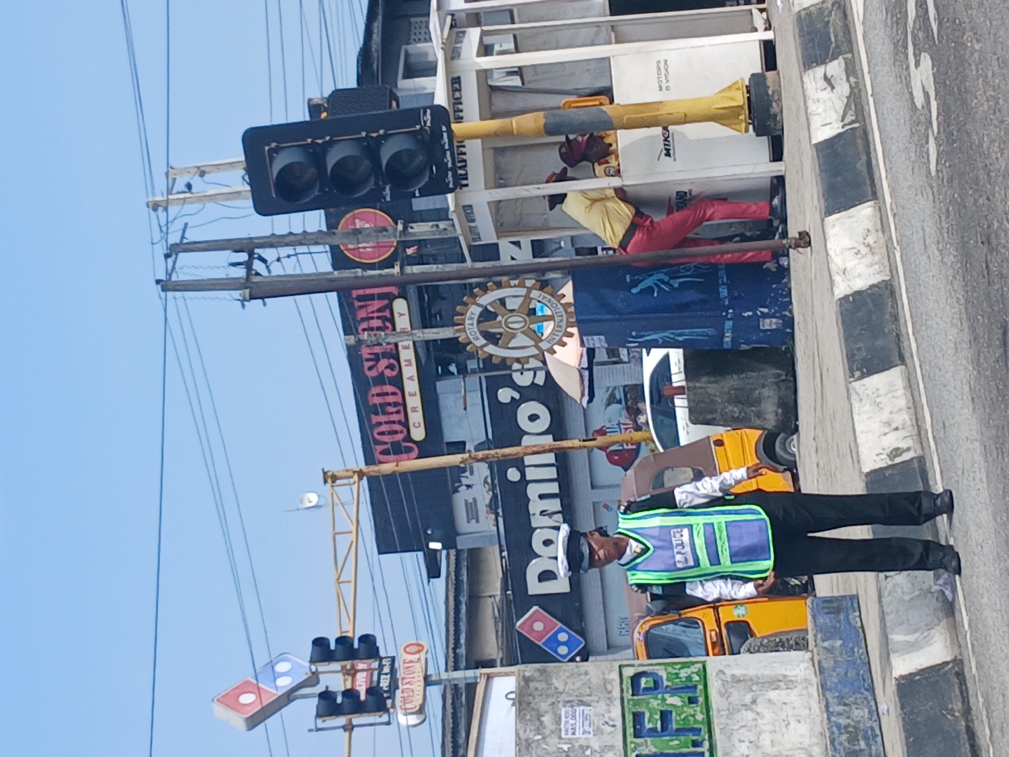 In Animashawun, Nigeria, a traffic officer stands on a street, directing traffic. The officer is wearing a dark uniform with a bright green and blue vest that says "POLICE." Behind the officer, a yellow auto-rickshaw is parked. In the background, there are several businesses, including a Domino's Pizza and a Cold Stone Creamery. Traffic lights are visible, and power lines crisscross the sky. A white booth with the words "TRAFFIC OFFICER" is also present, with two individuals inside.