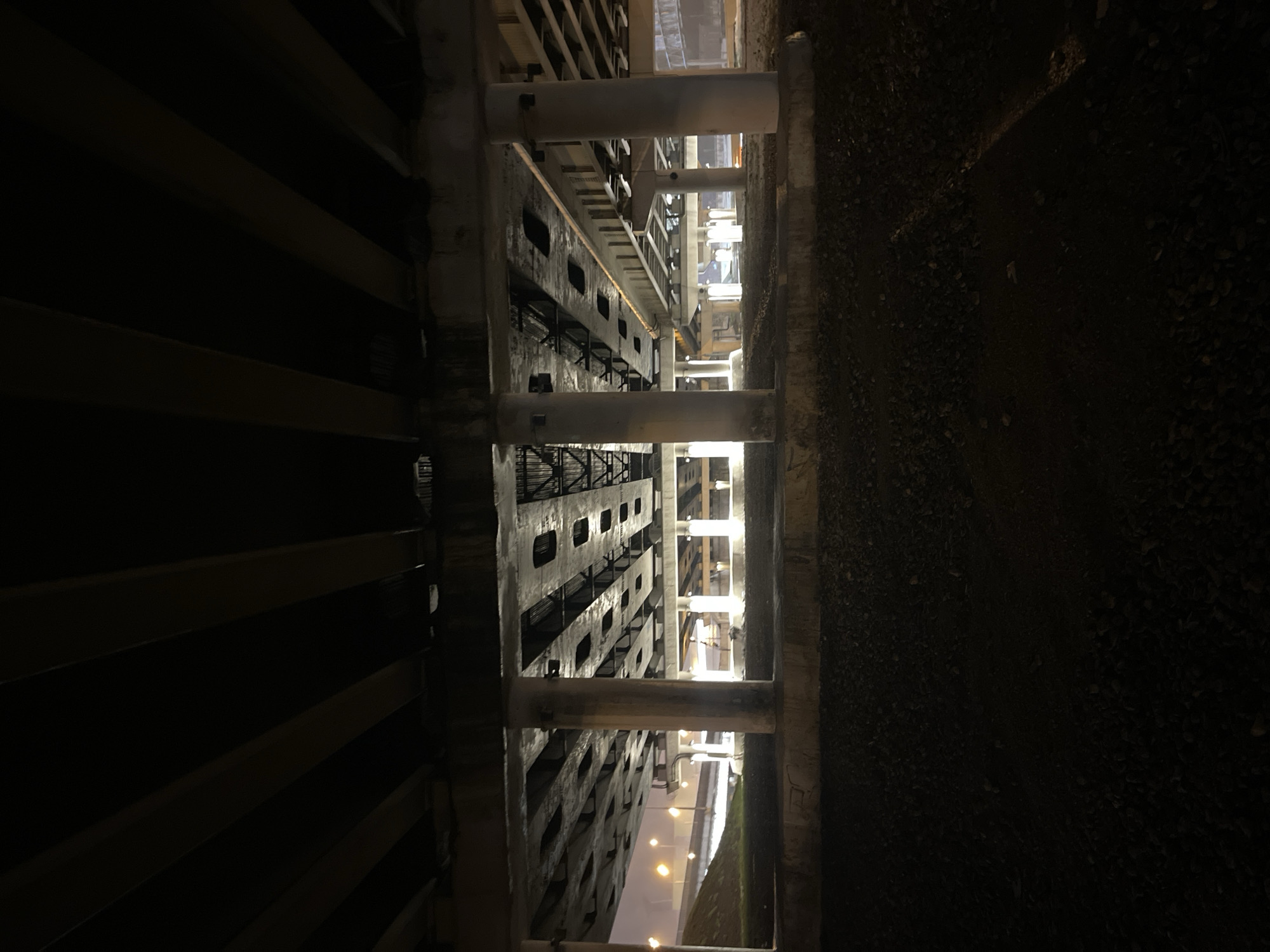 This nocturnal image captures a dimly lit, stark industrial or utility area, likely in Khimki, Russia. The scene is dominated by concrete and metal structures, giving it a functional and somewhat desolate atmosphere.
On the left side, a large, dark building or barrier features numerous prominent horizontal beams or slats, creating deep shadows and a repetitive pattern. These elements suggest a robust, possibly multi-story, structure like a warehouse facade or part of a large loading dock.
In the center, a passage or alley separates this structure from another. Within this gap, a multi-story building is visible, characterized by a grid-like pattern of rectangular openings, which could be windows, ventilation shafts, or structural voids. Several thick, light-colored concrete pillars stand vertically, traversing the space and likely supporting an overhead section or a bridge. These pillars, along with other visible structural elements, are intermittently lit by artificial light sources, casting a glow on their surfaces.
To the right, the ground is dark and uneven, appearing to be covered in gravel or rough terrain, receding into the darkness. The overall lighting is sparse, primarily from distant or hidden artificial sources, emphasizing the architectural forms and the deep shadows. There is no visible human activity or discernible text in the image. The time of day is clearly night.