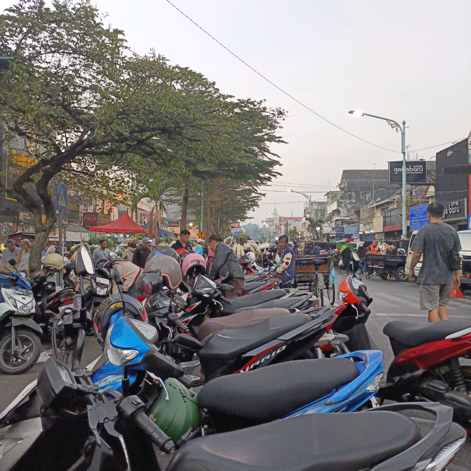 The image depicts a street scene in Salatiga, Indonesia, likely a marketplace or commercial area. The foreground is dominated by a large number of parked motorcycles, many with helmets placed on them. In the background, a street is lined with various shops and stalls, some with awnings and signage. A prominent red tent-like structure is visible, possibly a vendor's stall. Several people are seen walking and interacting in the middle ground, some appearing to be customers or vendors. A street lamp with decorative elements is lit, indicating it is either dusk or a dimly lit area. Trees line the street, contributing to the urban environment. The overall atmosphere appears to be that of a busy, active commercial district.