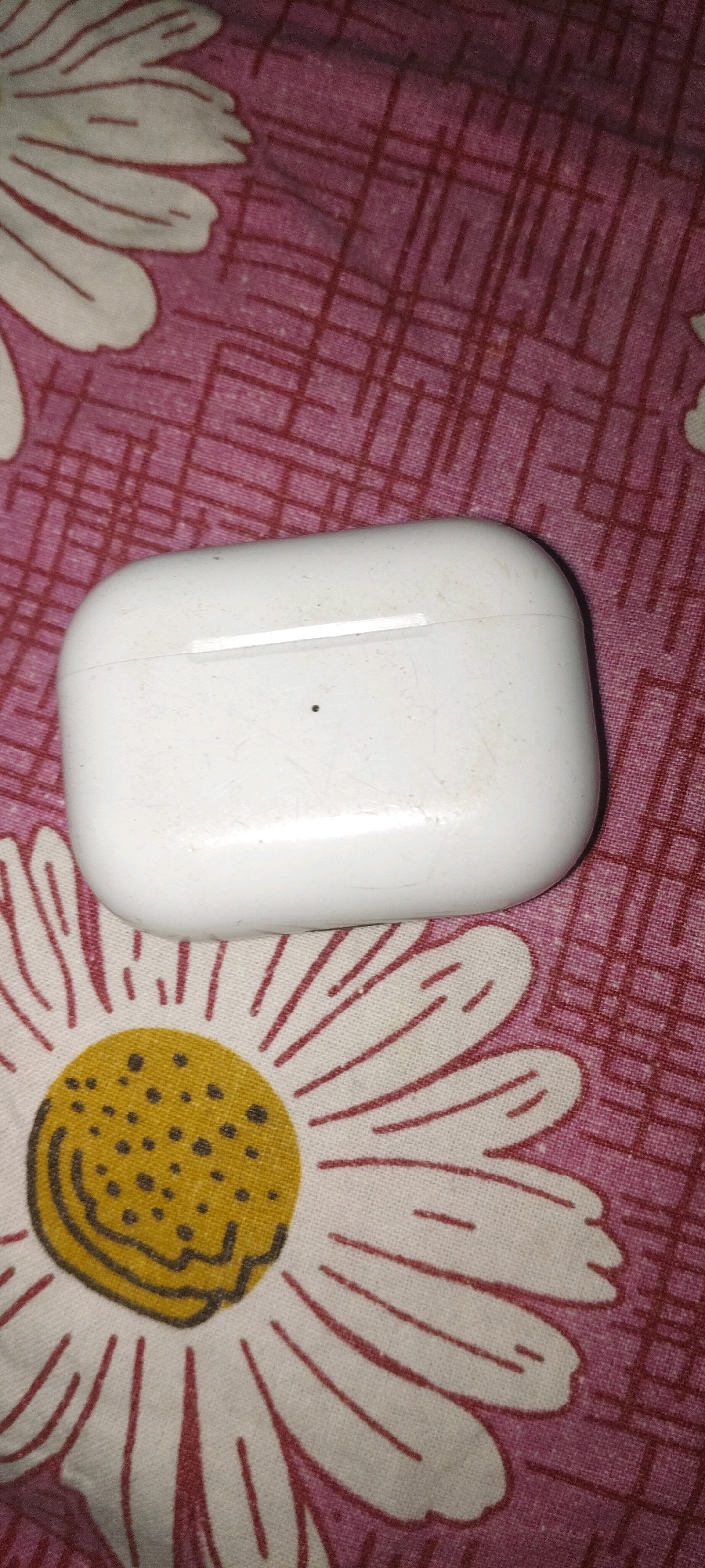 A white wireless earbud charging case rests on a pink and white floral patterned fabric. The fabric has a repeating pattern of white daisy-like flowers with yellow centers and red stems and petals. The charging case is positioned in the center of the image, slightly tilted. It appears to be closed and has a small black dot on its surface. The fabric has some visible texture and creases. The scene is indoors, likely in a domestic setting, and there are no people visible. The specific location in Appanapalle, India, is not discernible from the image alone.