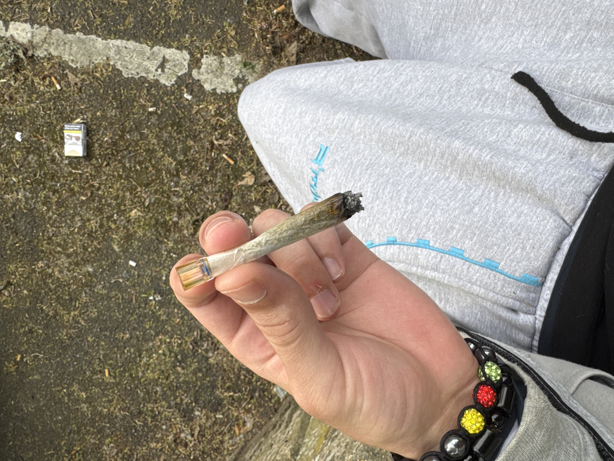 A person is holding a lit joint in their right hand. The person is wearing gray sweatpants with a turquoise design on the left thigh, a black drawstring at the waist, and a black-beaded bracelet with multi-colored, rhinestone-studded beads on their wrist. The background includes a cigarette pack and cigarette butts on what appears to be a patch of ground near a road. The location is in the city of Les Ulis, France.