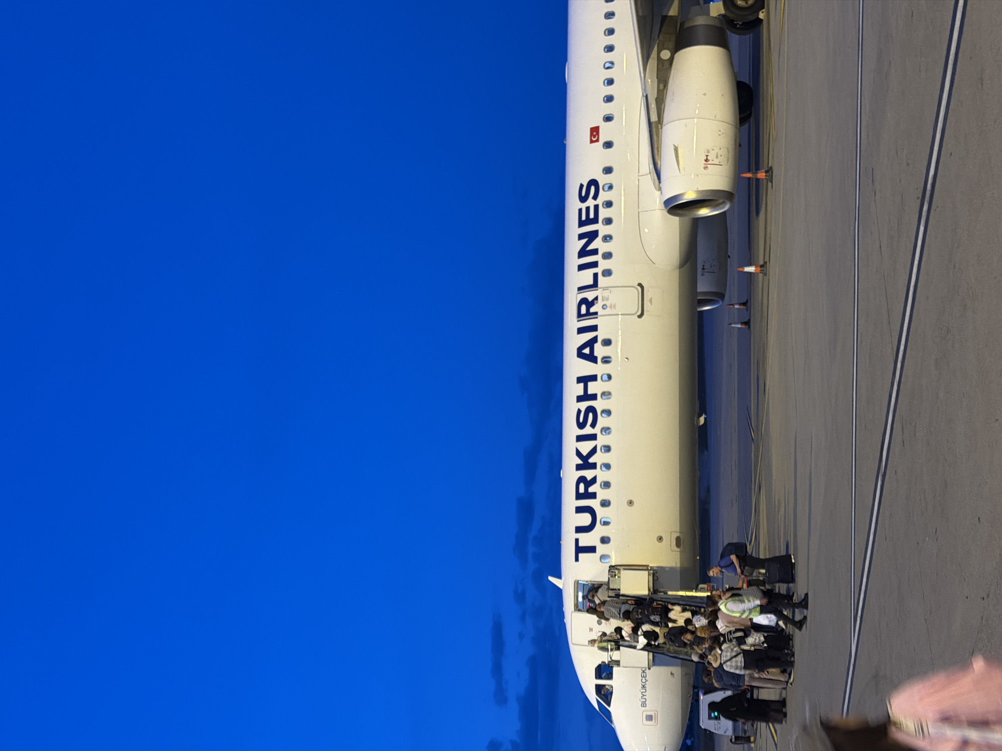 The image shows a Turkish Airlines plane on the ground, presumably at an airport. Passengers are disembarking using a mobile stairway. The plane's fuselage is visible, with the airline's name prominently displayed. The sky is a clear blue in the background. There are markings on the tarmac near the aircraft and people on the ground waiting to board or assisting passengers. The text on the airplane reads "Büyükçeki." There is no information present to suggest the location as Podgorica, Montenegro. The image does not reveal any details that confirm a specific city location.