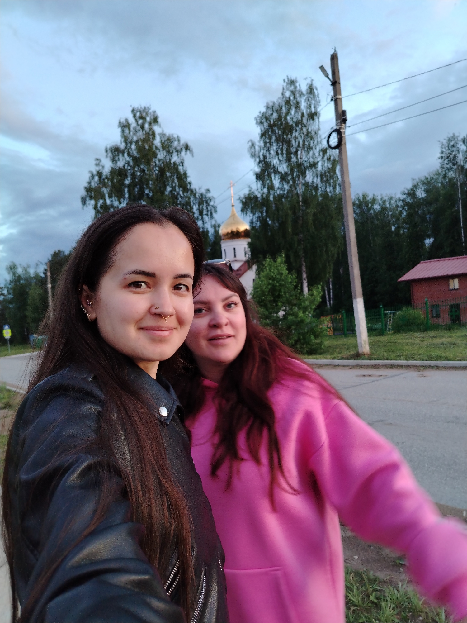 Two women stand side-by-side on a paved surface, likely a sidewalk or road, with a street visible to the side. The woman on the left wears a black leather jacket and has dark, long hair with piercings on her ear and nose. The woman on the right is wearing a pink hoodie and has reddish-brown hair.
In the background, there's a building with a golden dome, likely a Russian Orthodox church, positioned behind a line of green trees. A utility pole stands tall, with wires stretching from it. Behind the women, across a grassy area, is a small red brick building. The sky is overcast.
The image was taken in Noviy, Russia.