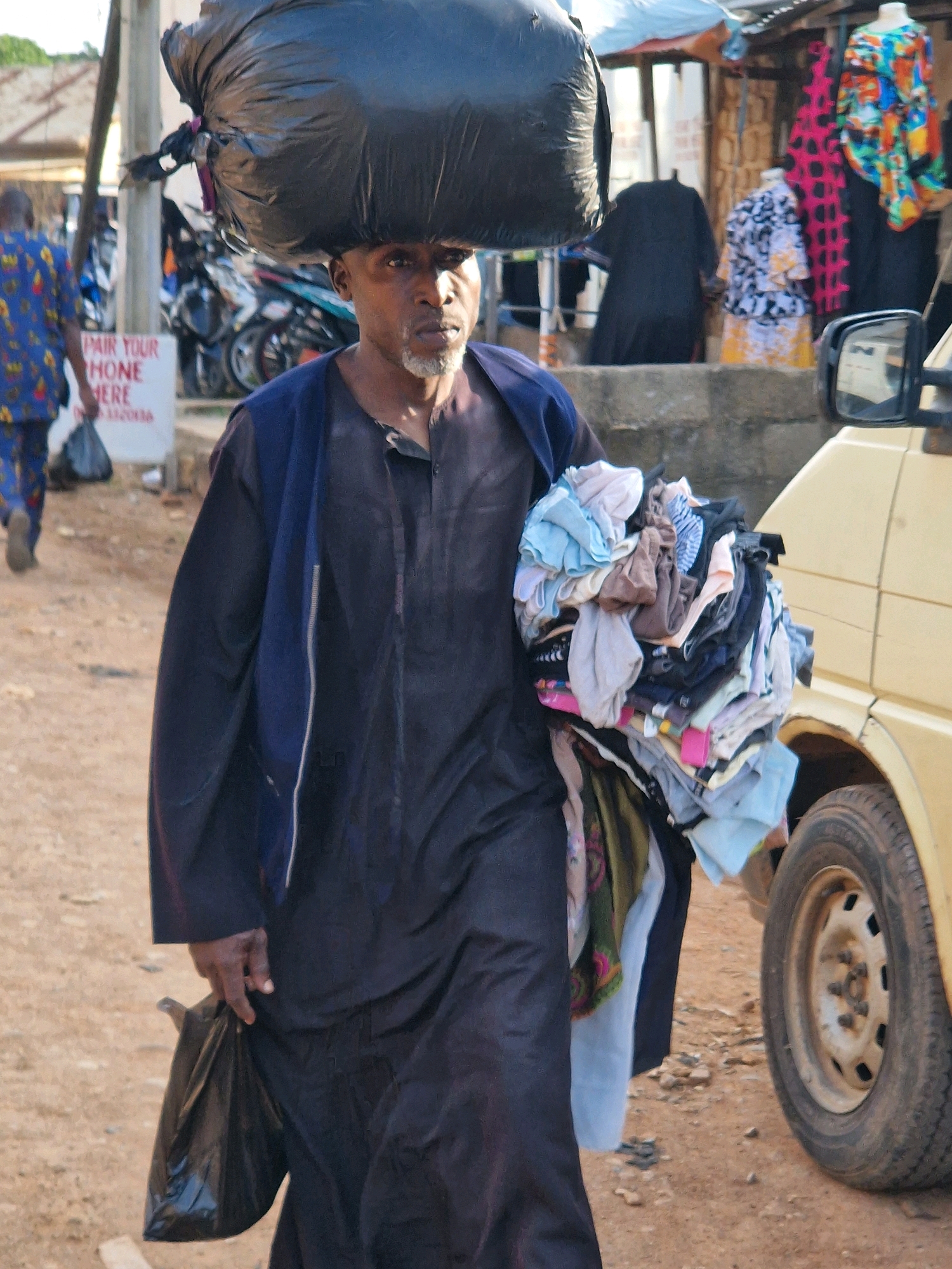 The image shows a man walking outdoors, carrying a large black trash bag on his head and a bundle of clothes in his left arm. He is wearing a dark robe and has a gray beard. To his right, a portion of a yellow vehicle is visible, including its front wheel and side mirror. In the background, there is a market stall with various colorful clothing items displayed on mannequins. A person wearing a blue and yellow patterned shirt is walking away from the viewer on the left side of the frame, carrying a black bag. The ground appears to be dirt and gravel.