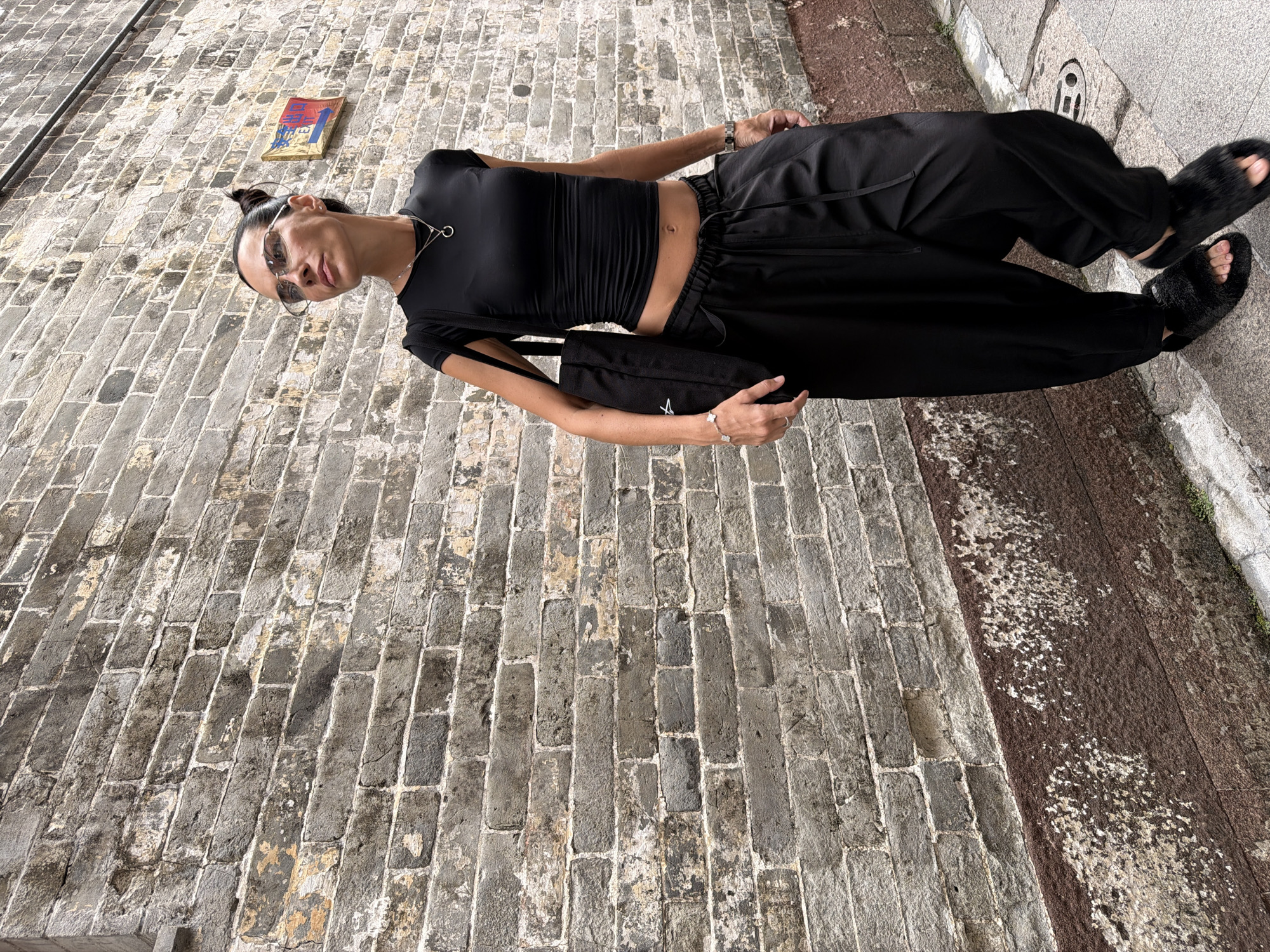 A woman with dark hair pulled into a bun walks down a narrow alleyway with a brick wall on her left. She is wearing a black cropped t-shirt, black wide-leg pants, and black furry slides. She carries a black tote bag over her right shoulder and wears silver sunglasses and a silver necklace. A small red and blue sign with Chinese characters is visible on the brick wall behind her. The ground is paved with gray stones, and a metal grate is visible in the foreground. The brick wall is old and weathered, with sections of missing mortar and discolored bricks. The overall scene suggests an urban setting in Chancheng District, China, possibly an older neighborhood due to the architecture and materials.
