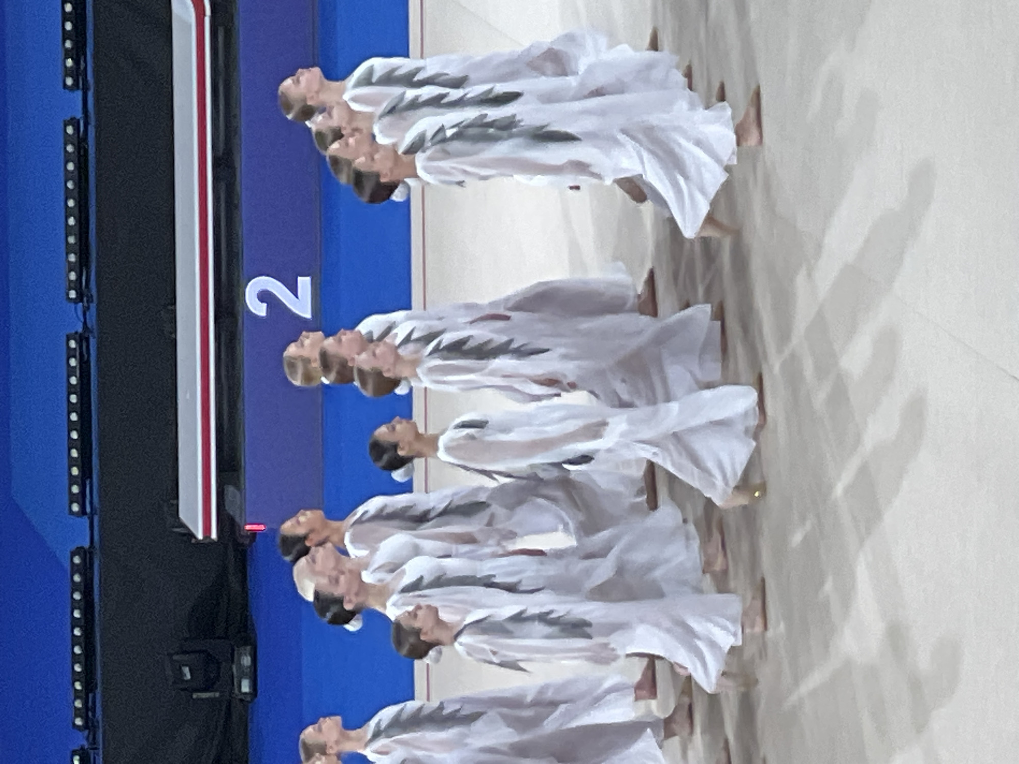 The image captures a group of young female gymnasts in white, flowing costumes with grey feather-like embellishments on the front, performing on a light-colored floor. They are arranged in a line, moving from left to right across the frame. The background features a dark blue wall with a prominent white numeral "2" and rows of lights above. The lighting suggests an indoor arena setting. The scene appears to be a competition or performance, possibly a synchronized routine given the uniformity of the outfits and the coordinated movement. The image quality is somewhat grainy, indicating it was taken with a mobile device or in low light conditions. The specific location in Moscow, Russia cannot be determined from the visual information provided.
