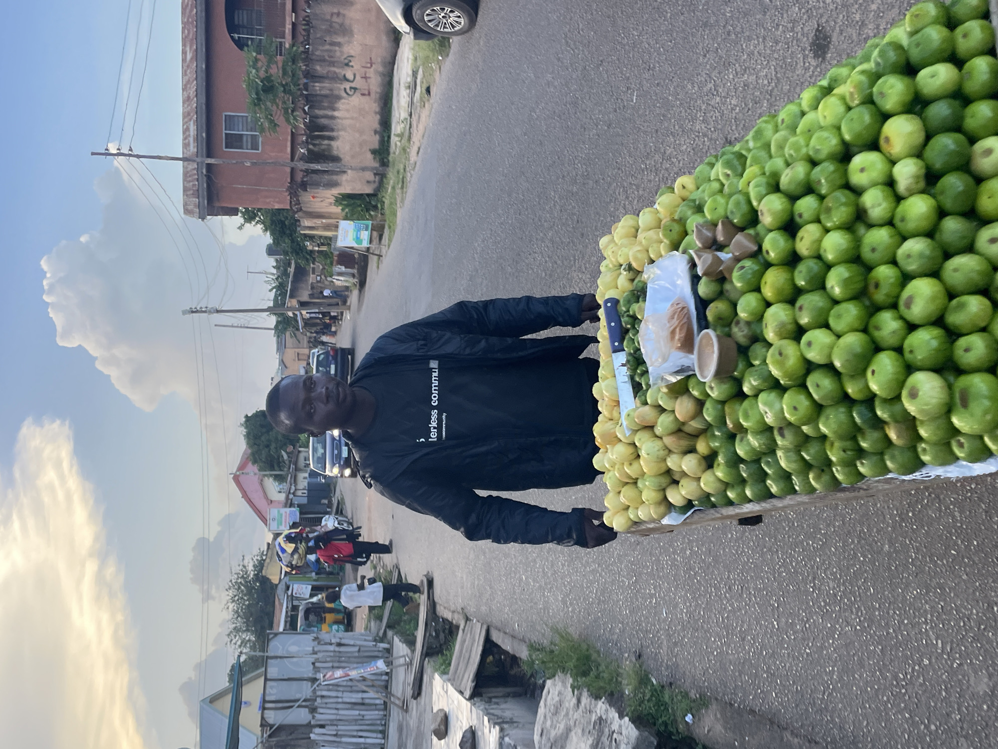 An outdoor medium shot captures a Black male vendor pushing a cart laden with produce down a paved road in Jos, Nigeria. The vendor, wearing a black long-sleeved shirt with white text that reads "Terlerless Community," is positioned in the right foreground, facing slightly left and looking toward the viewer. His hands are on the cart's handle. The cart is overflowing with green fruits, with a smaller quantity of yellowish-green fruits on top. A knife and a small container of spices are visible on the cart.
The road extends into the background, flanked by buildings and vegetation. To the left of the road, a shallow ditch runs parallel to it, spanned by wooden planks. Several other people and vehicles are visible in the background, suggesting a populated urban environment. A yellow motorized tricycle is parked on the left side of the road, with a person in a red shirt carrying items on their head walking away from it. A light-skinned person in a white shirt and dark pants is walking further down the road on the left. A dark-colored car is parked on the right side of the road. The sky above is partly cloudy with dramatic cloud formations illuminated by soft sunlight. Buildings in the background include a two-story structure with a reddish-brown facade and another building with a light-colored exterior. Graffiti is visible on a wall.