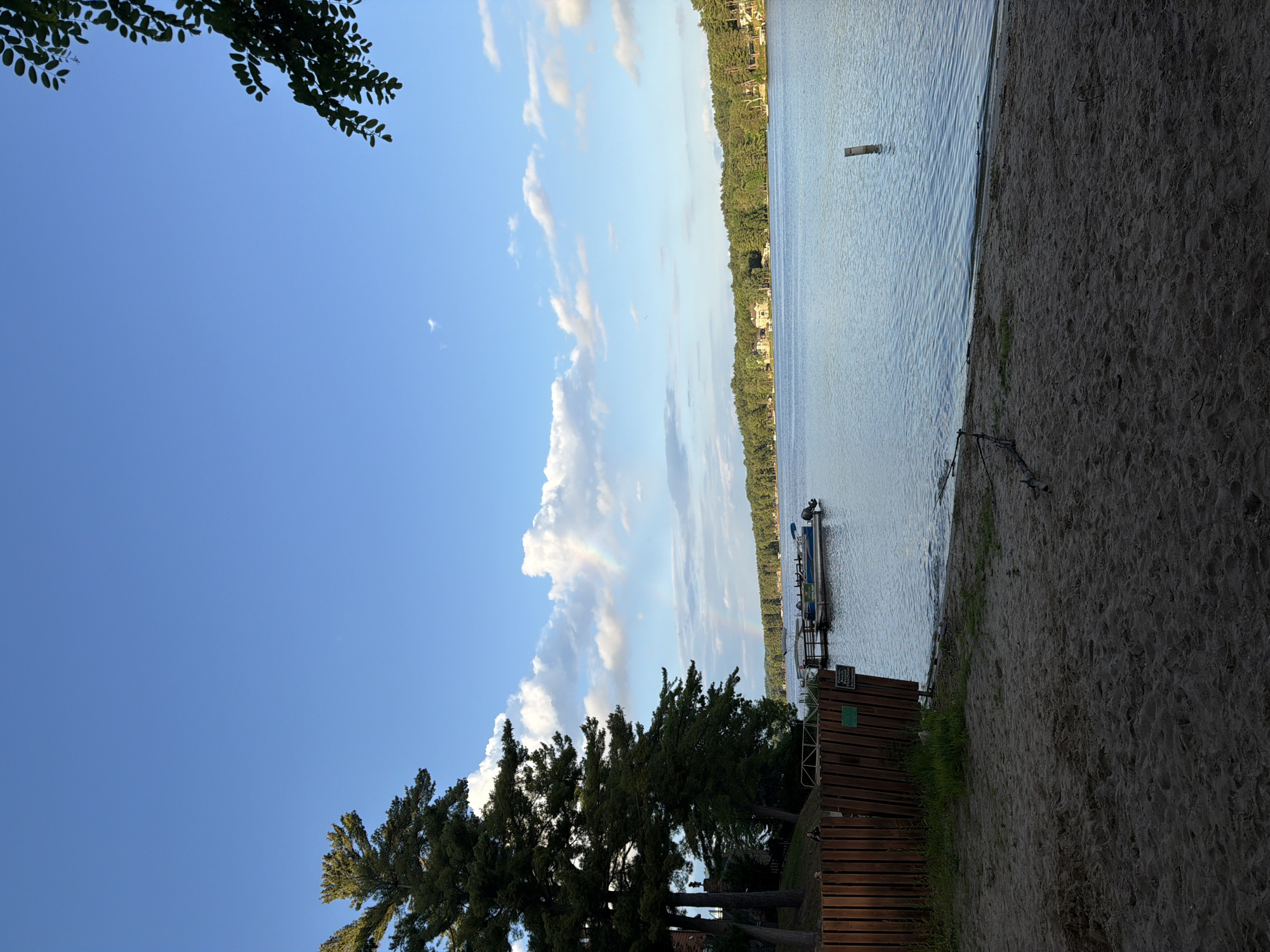 The image presents a detailed lakeside scene captured during daylight hours in Lake Delton, United States of America.
The upper portion of the frame is dominated by a clear blue sky, interspersed with scattered, puffy white cumulus clouds. A distinct, albeit faint, rainbow arc is visible in the upper-mid-left section of the sky, appearing to originate from above the distant treeline.
On the left side, a cluster of tall, dark green coniferous trees, possibly pines, extends from the bottom of the frame towards the upper-left, partially obscuring a building structure visible beneath their foliage. A sturdy wooden fence, constructed from vertical planks, runs along the left foreground, parallel to the water's edge. Attached to the fence is a small green sign with white text, which is largely illegible. Behind the fence, nestled near the trees, a solitary individual with dark hair and clothing is partially visible, facing towards the lake.
The central and mid-ground is occupied by a calm body of water, likely a lake, exhibiting minor ripples on its surface and reflecting a soft blue-grey hue. A wooden dock extends from the left shore into the water, with two pontoon boats moored to it. The closer boat is blue and white with an outboard motor visible at the stern. The second boat, positioned further back along the dock, appears lighter in color, possibly white or light grey, and features a dark canopy. In the mid-right section of the water, a single dark wooden post protrudes vertically. The distant shoreline across the lake is densely forested with a mix of evergreen and deciduous trees, appearing as a dark green band. Several houses or residential structures with varying roof colors are visible nestled among the trees on the far shore.
The immediate foreground consists of a sandy beach, appearing darker in tone due to being in shadow. The sand displays visible undulations and slight depressions, suggesting footprints. Sparse patches of green grass are present near the water's edge on the sand. A piece of driftwood or a fallen branch lies horizontally on the sand in the lower-right corner. In the upper-right corner, a dark, leafy tree branch extends into the frame, partially silhouetted against the sky.