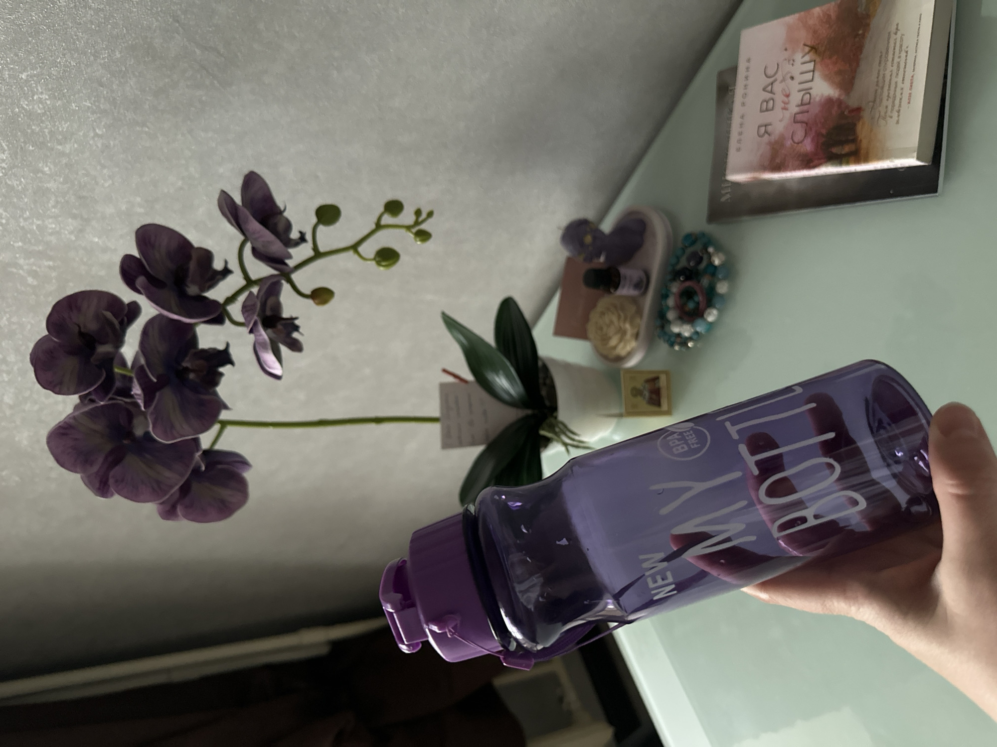 A hand is holding a purple water bottle with the words "NEW MY BOTTLE" printed on it in white. The bottle is partially transparent and contains a clear liquid. Behind the bottle, a purple orchid plant with several blooms and green leaves is visible. To the right of the orchid, there are several small decorative items on a table, including what appears to be a small candle in the shape of a person, a bottle of essential oil, a beaded bracelet, and a small framed image of a religious figure. Further to the right, two books are stacked on the table. One book is white with a pink cover featuring an illustration of a tree-lined path with pink trees and text that reads "Я ВАС не слышу". The table appears to be made of glass and is set against a textured, off-white wall. The lighting suggests an indoor environment, possibly a room. There is no indication of the city of Vologda, Russia, in the image.